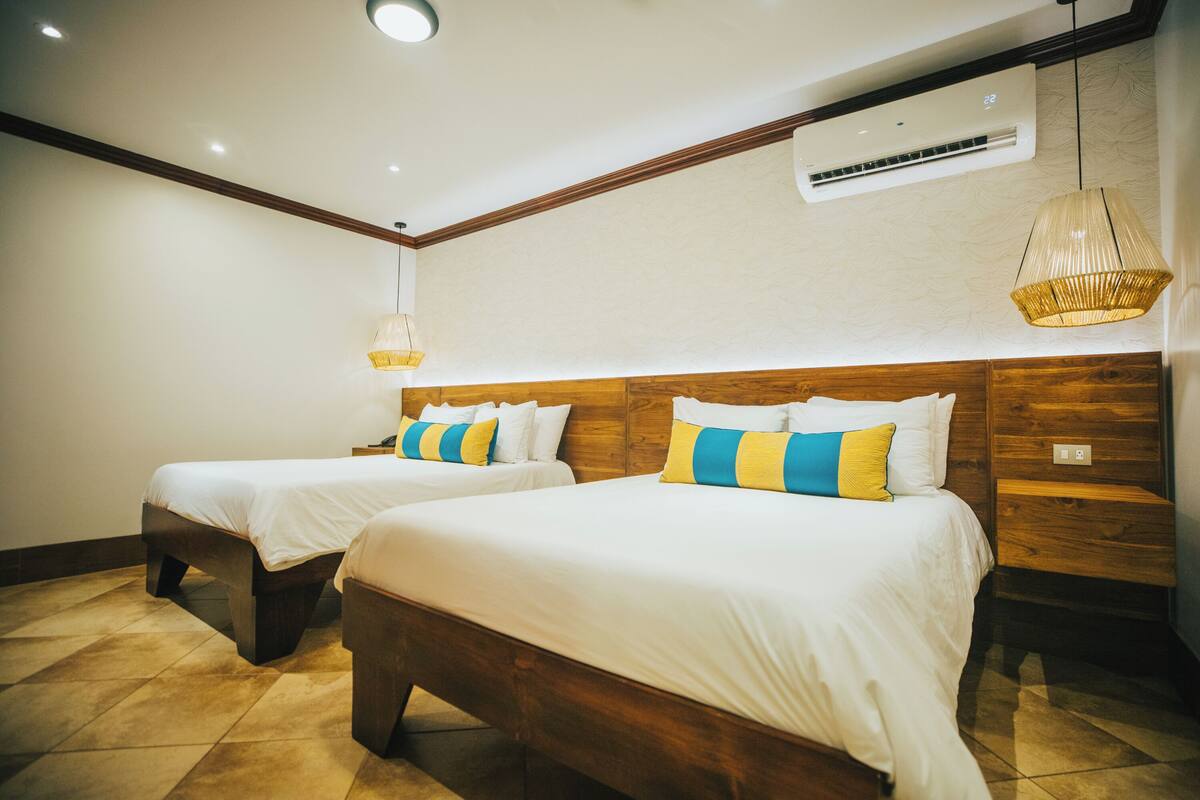 A hotel room with two beds, wooden headboards, a hanging light fixture, and an air conditioning unit.