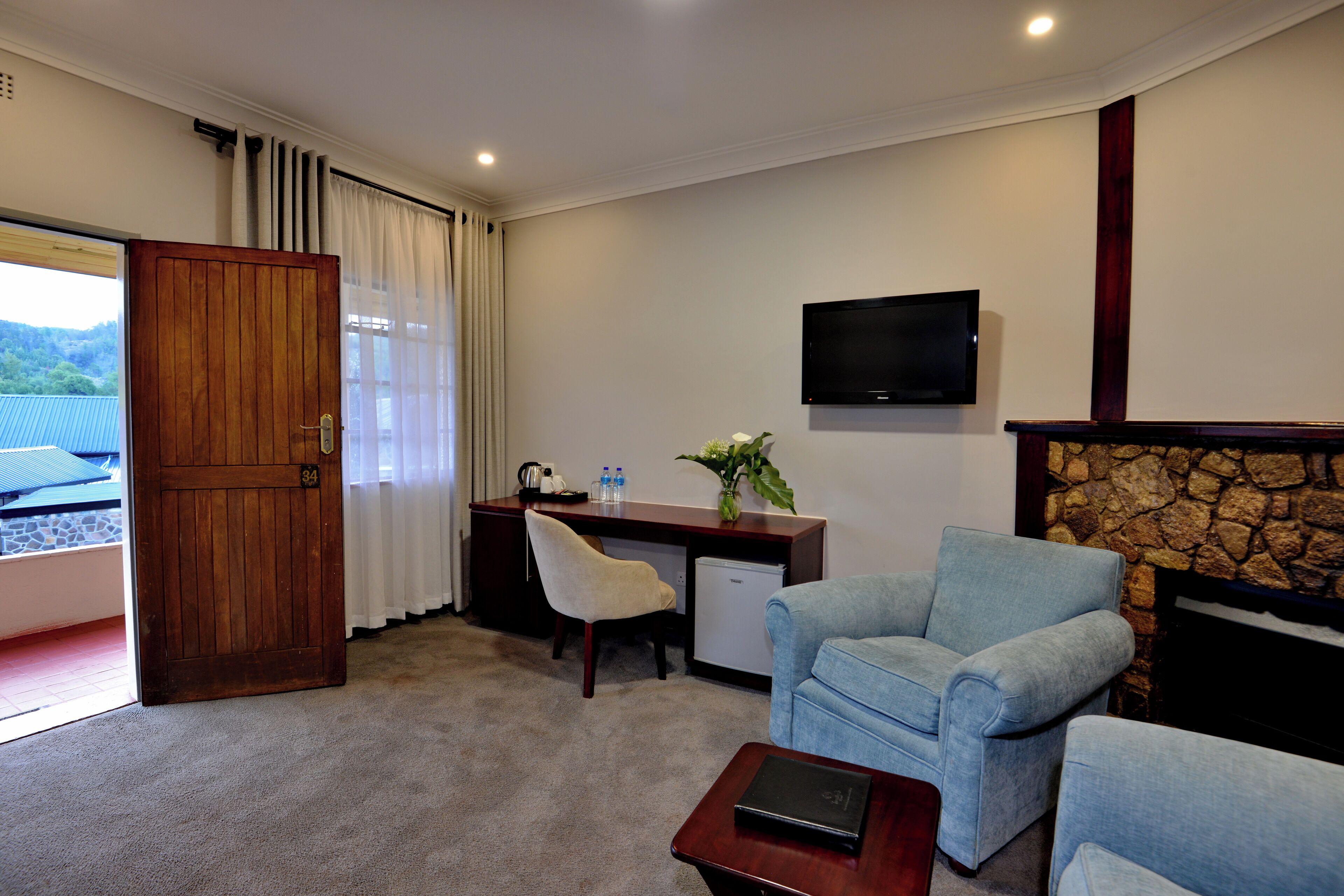 A hotel room with a wooden door, a desk with a chair, a blue armchair, a small TV, and a stone fireplace.