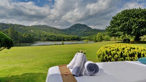 A bed with towels and a brown ribbon, overlooking a grassy field and a lake with hills in the background. - Troutbeck Resort (Nyanga)