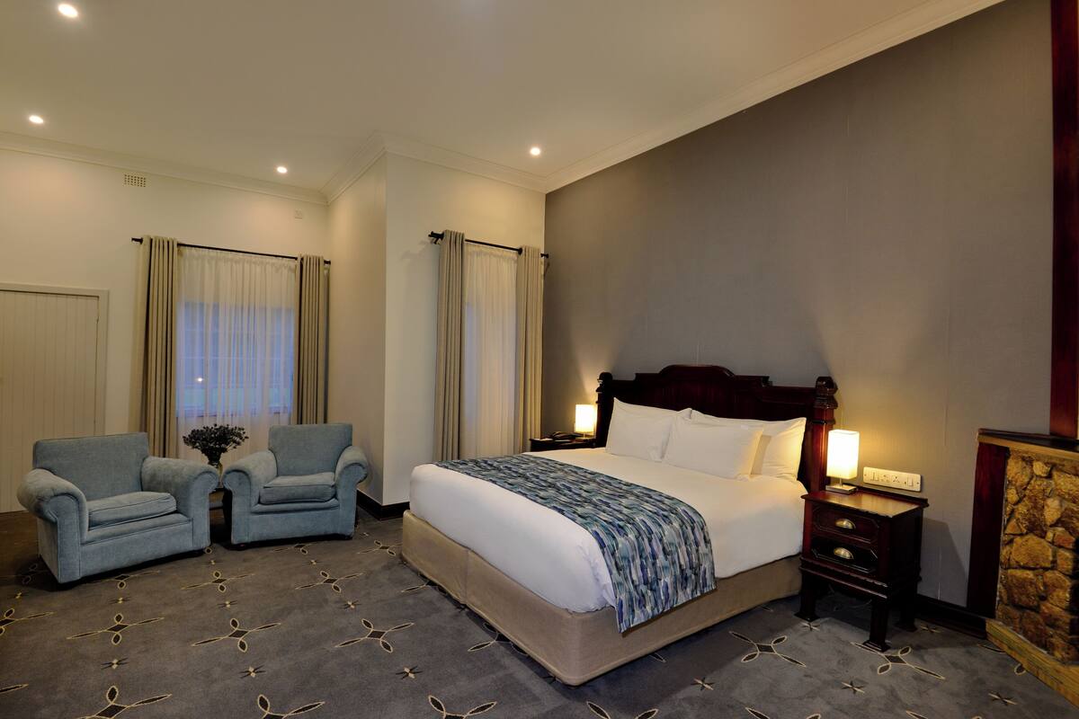 A hotel room with a large bed, two armchairs, a nightstand with a lamp, and a stone fireplace.