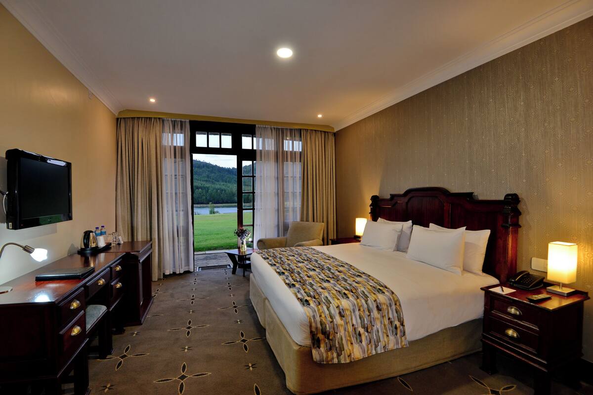 A hotel room with a large bed, a desk with a TV, a chair, and a view of a green landscape through the window.