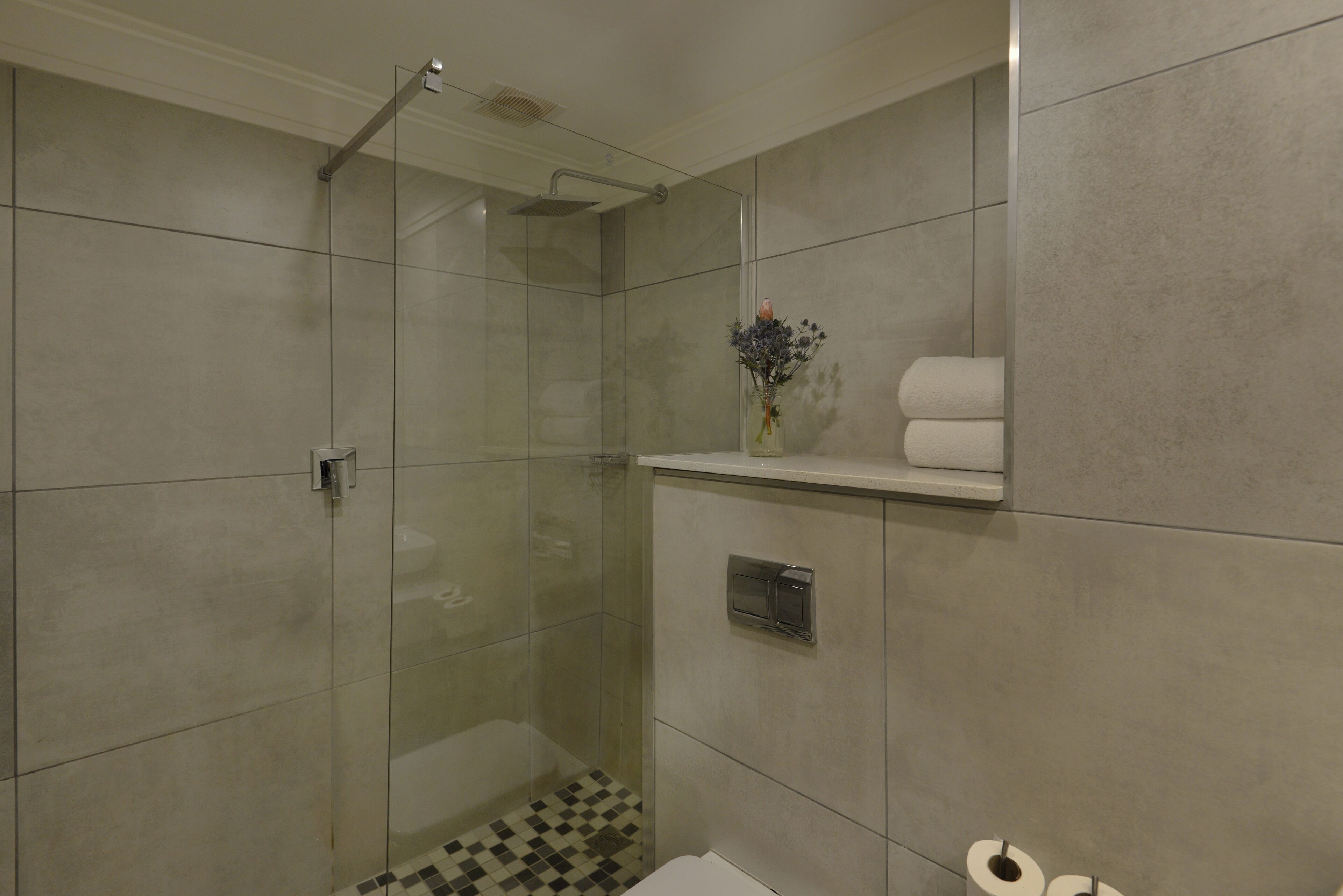 A modern bathroom with a glass-enclosed shower, a toilet paper holder, and a small shelf with towels.
