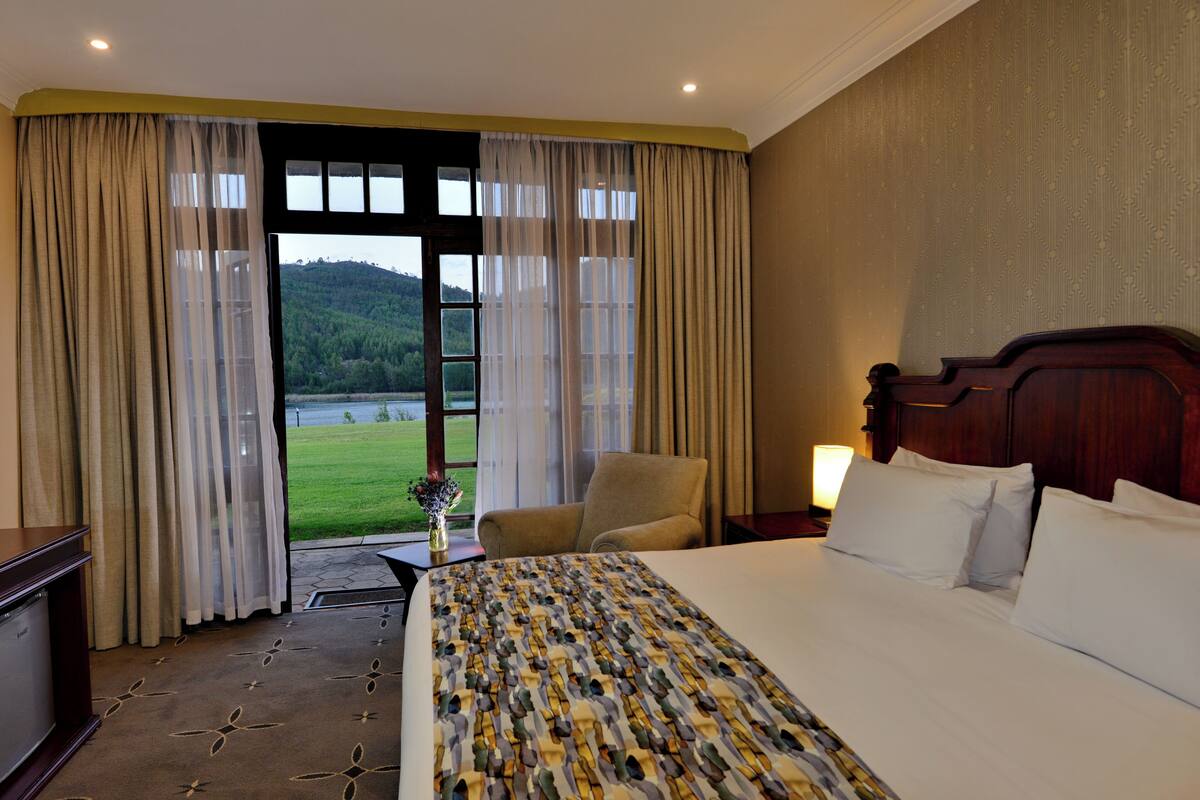 A hotel room with a large bed, a chair, a desk, and a view of a green landscape through the window.