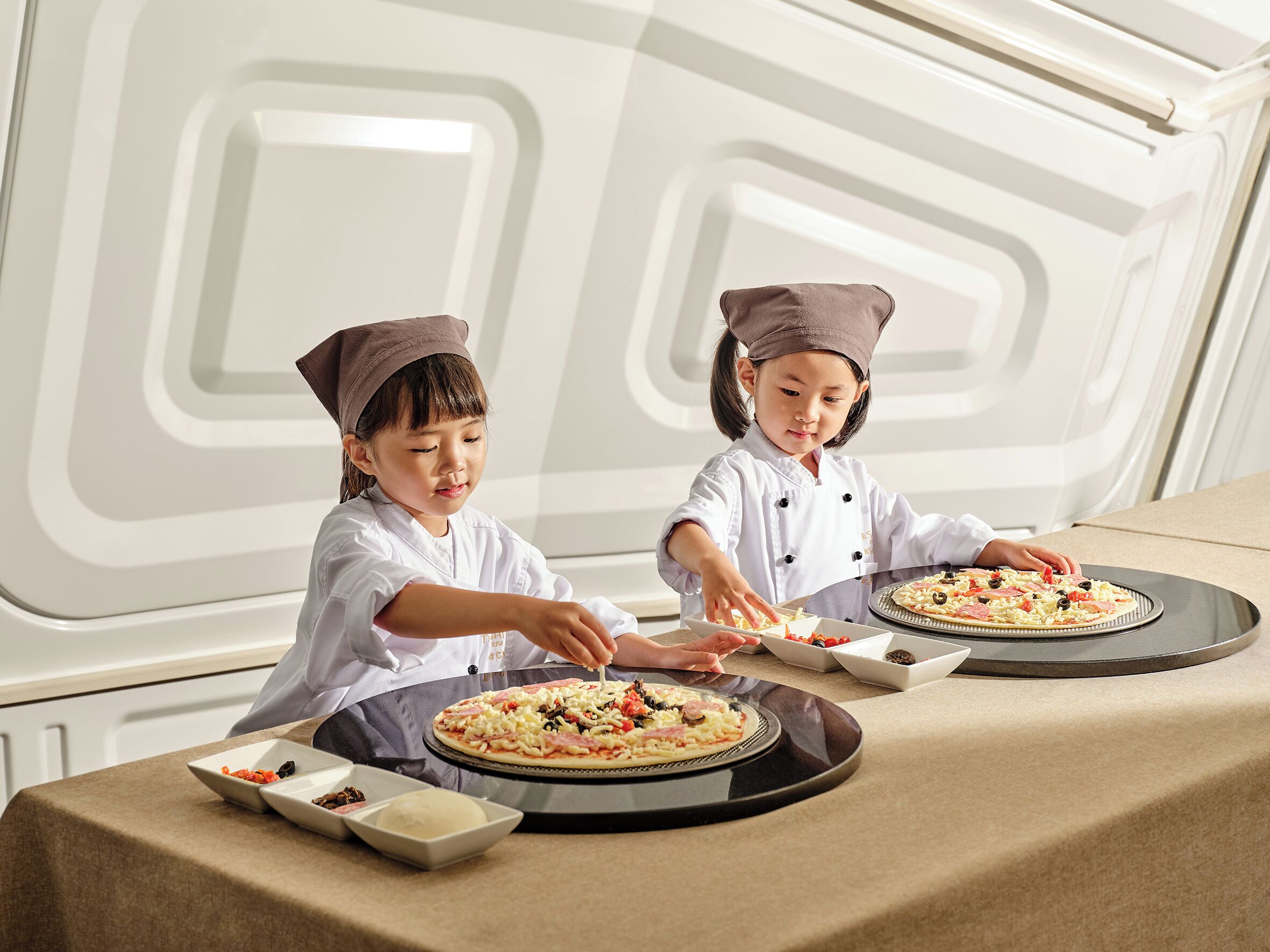 Deux enfants portant des toques de chef préparent des pizzas sur une table recouverte d’une nappe beige.