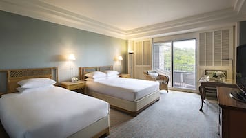 A hotel room with two beds, a TV, a desk, and a balcony with a view of greenery.