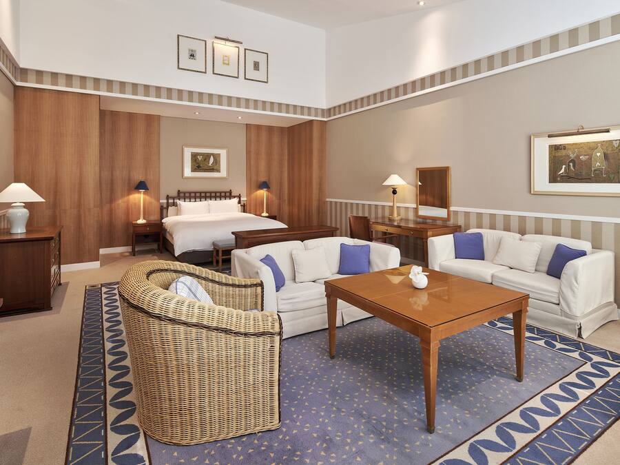 Een hotelkamer met een bed, twee witte banken, een rieten stoel, een houten tafel en een kamerkleed met een patroon.