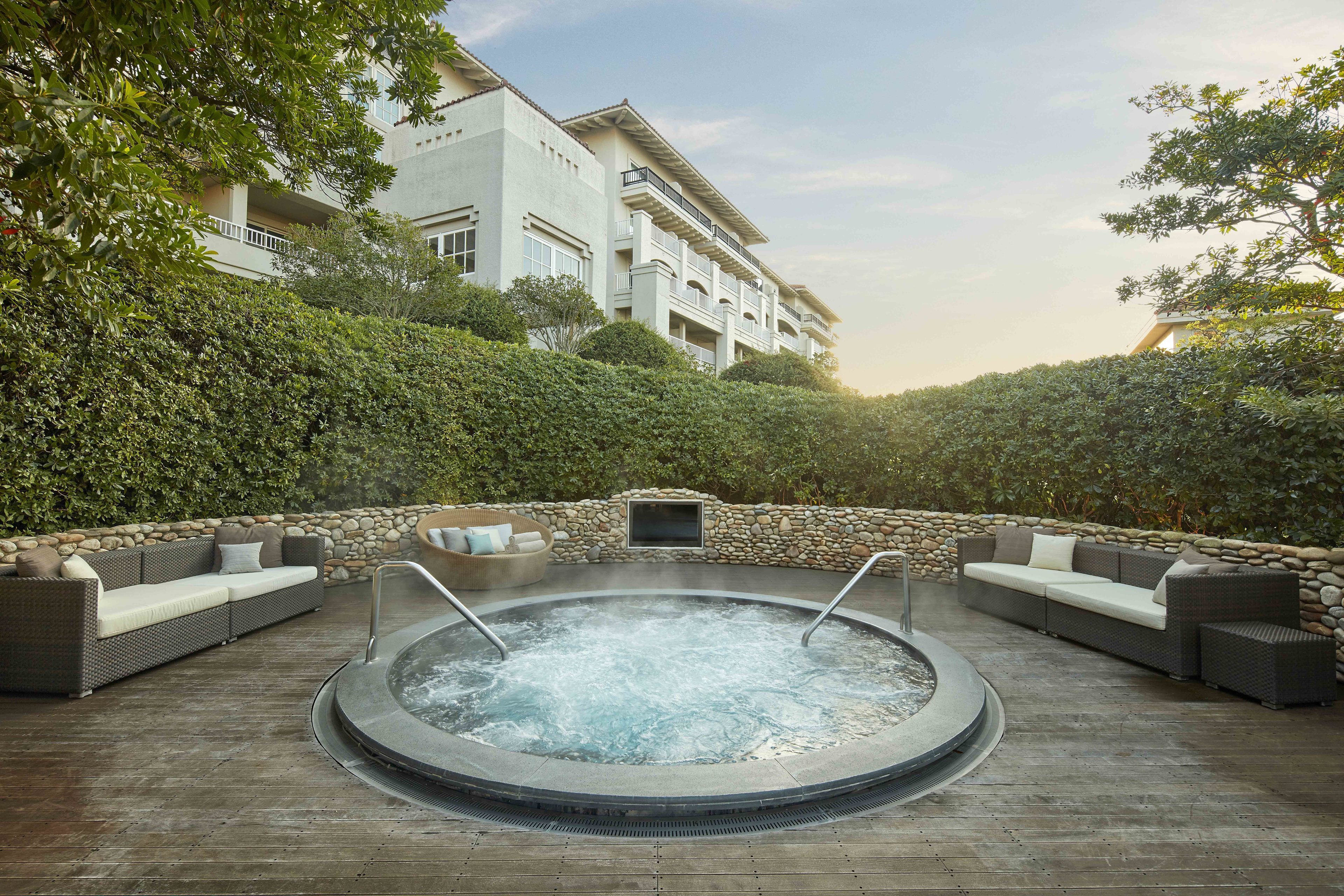 A hot tub surrounded by outdoor seating, a stone wall, and lush greenery.