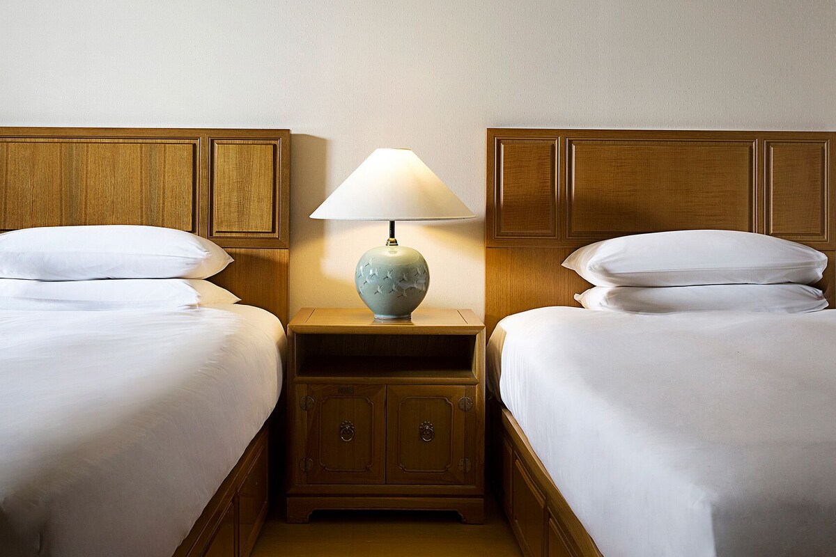 A hotel room with two single beds, a nightstand with a lamp, and wooden paneling on the walls.