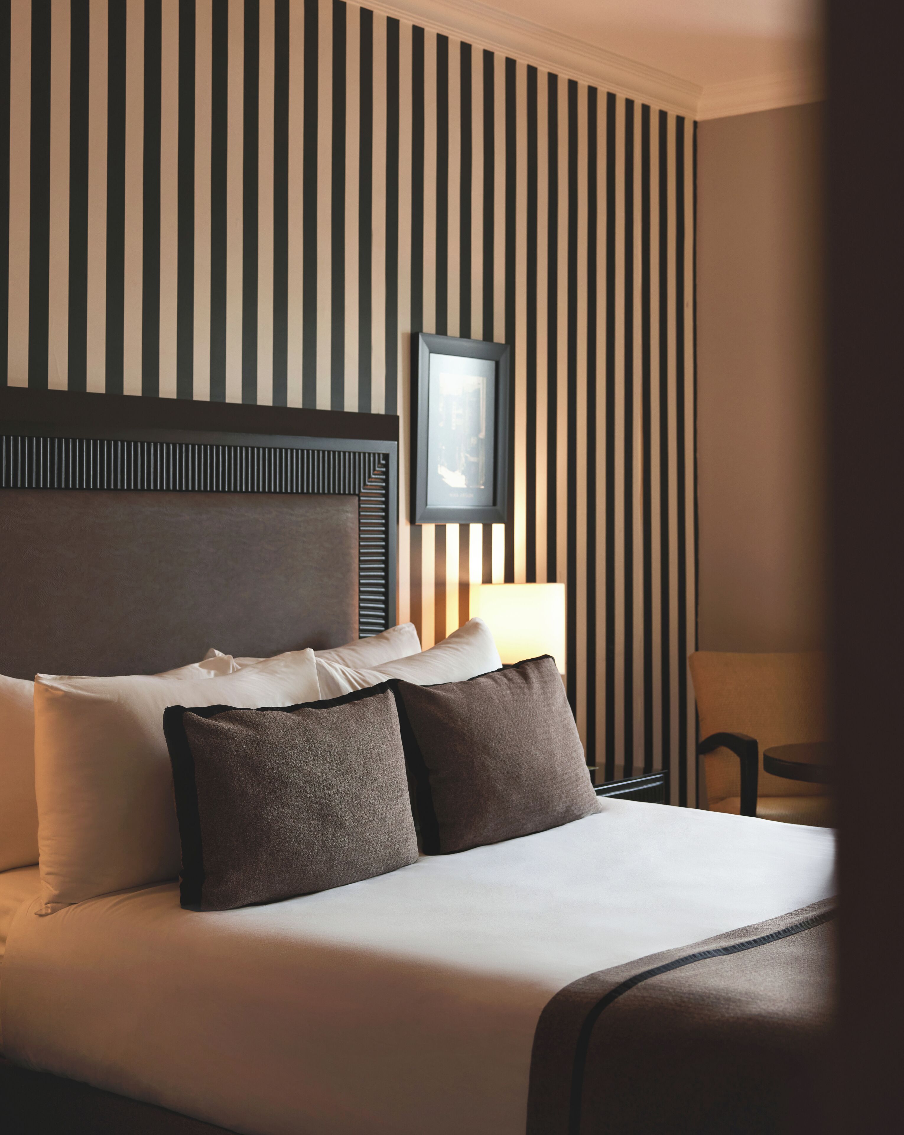 A hotel room with a large bed, a headboard, a bedside table, a chair, and a wall with vertical stripes.