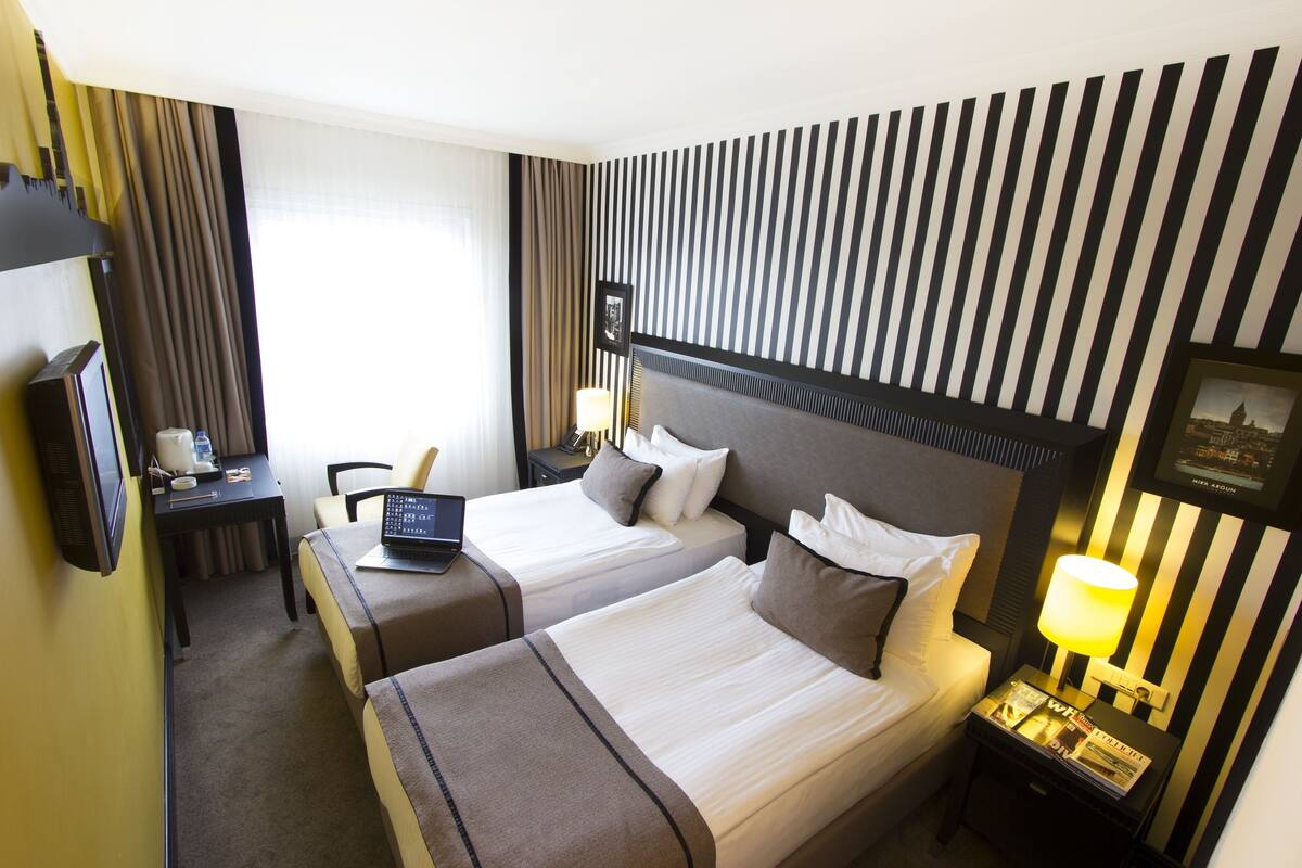 A hotel room with two beds, a desk with a laptop, a TV, and a nightstand with a lamp.