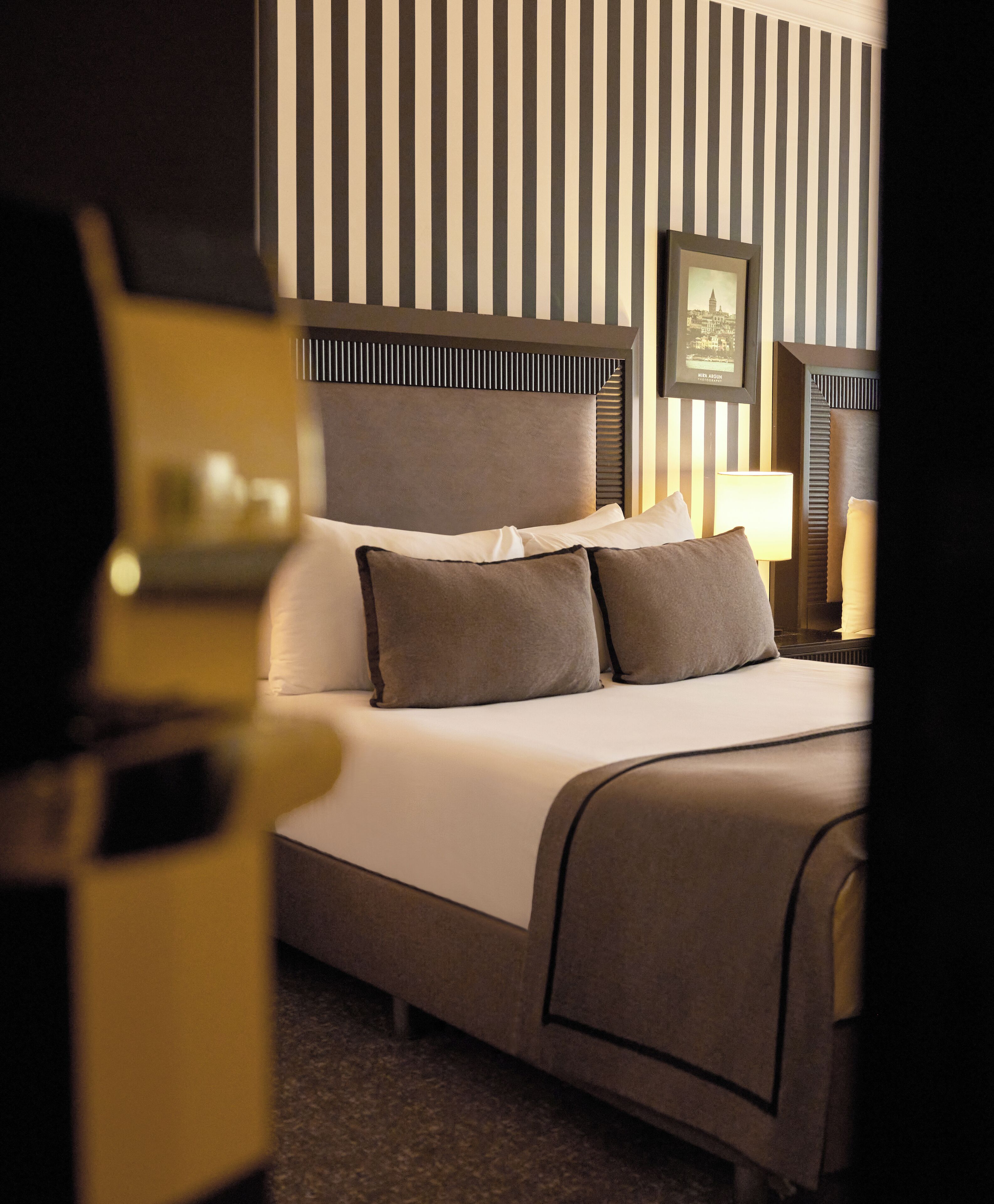 A hotel room with a large bed, a headboard with vertical stripes, a framed picture on the wall, and a bedside table with a lamp.