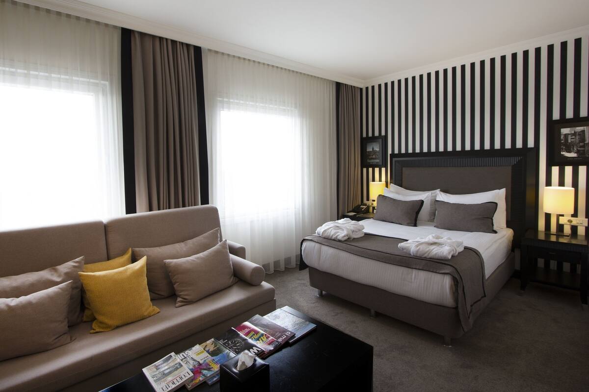 A hotel room with a bed, a sofa, a coffee table with magazines, and a nightstand with a lamp.
