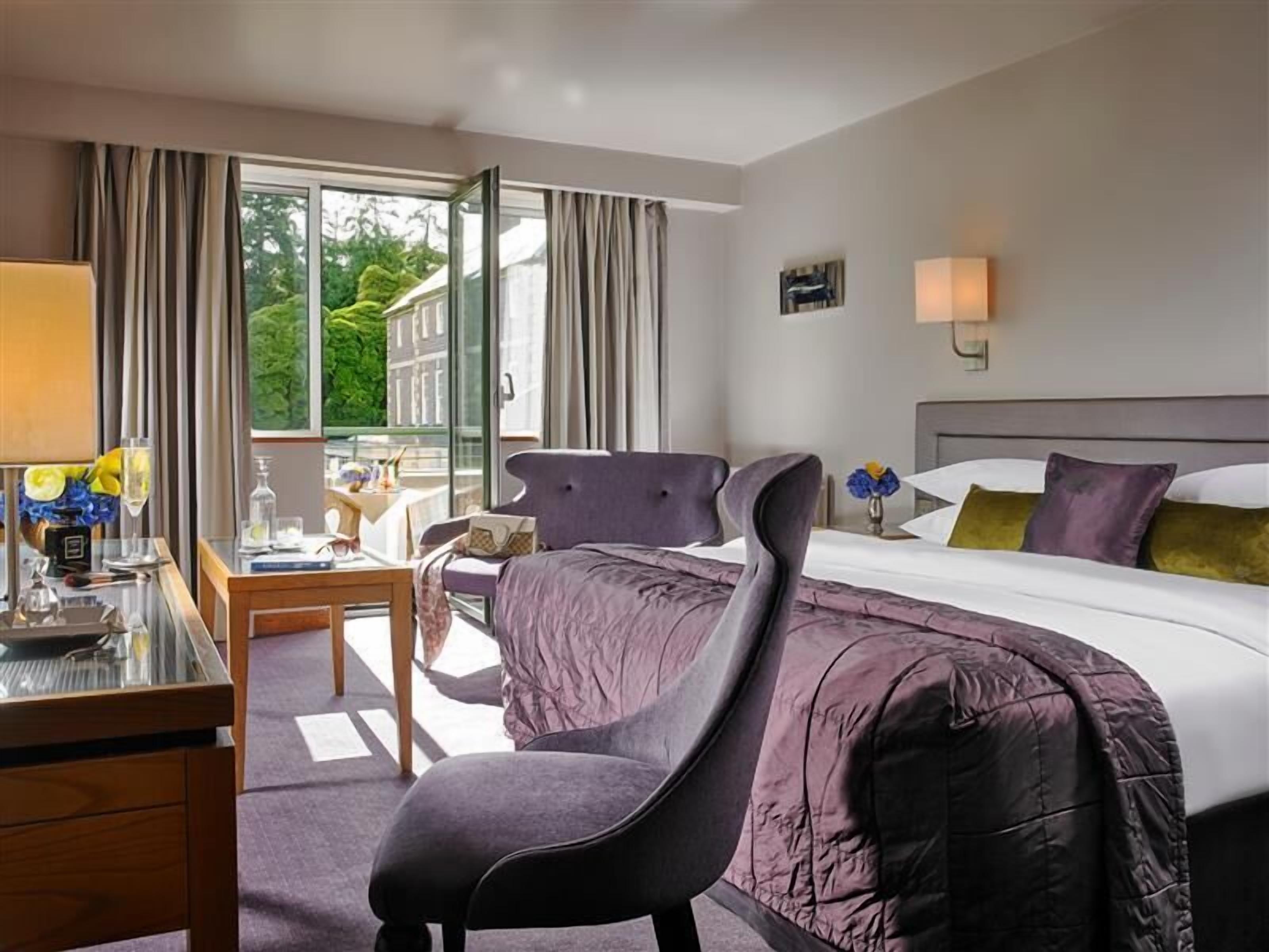 A hotel room with a bed, a desk, a chair, a window with curtains, and a view of greenery outside.