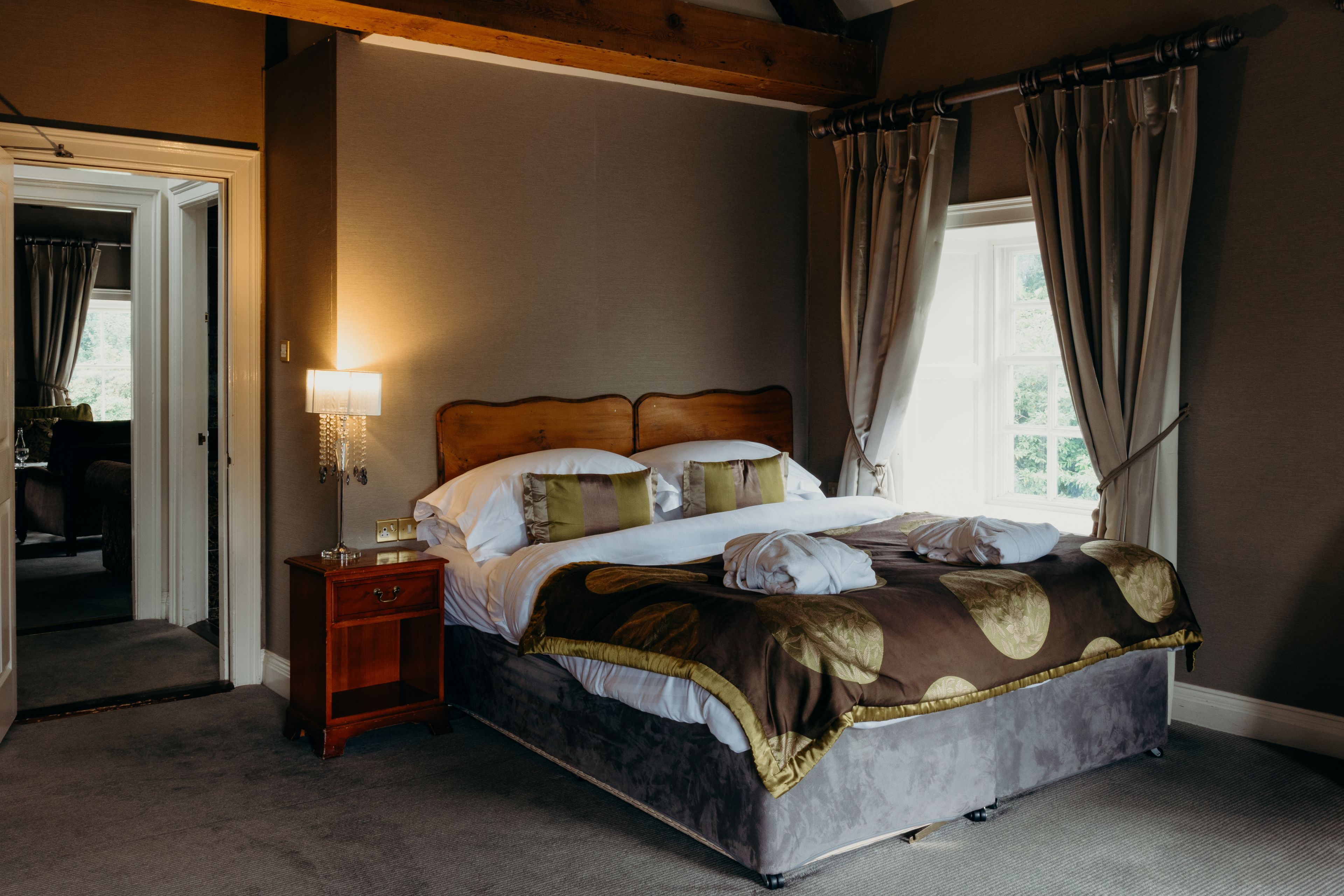 A bedroom with a wooden headboard, a bedside table with a lamp, a bed with a patterned bedspread, and a window with curtains.