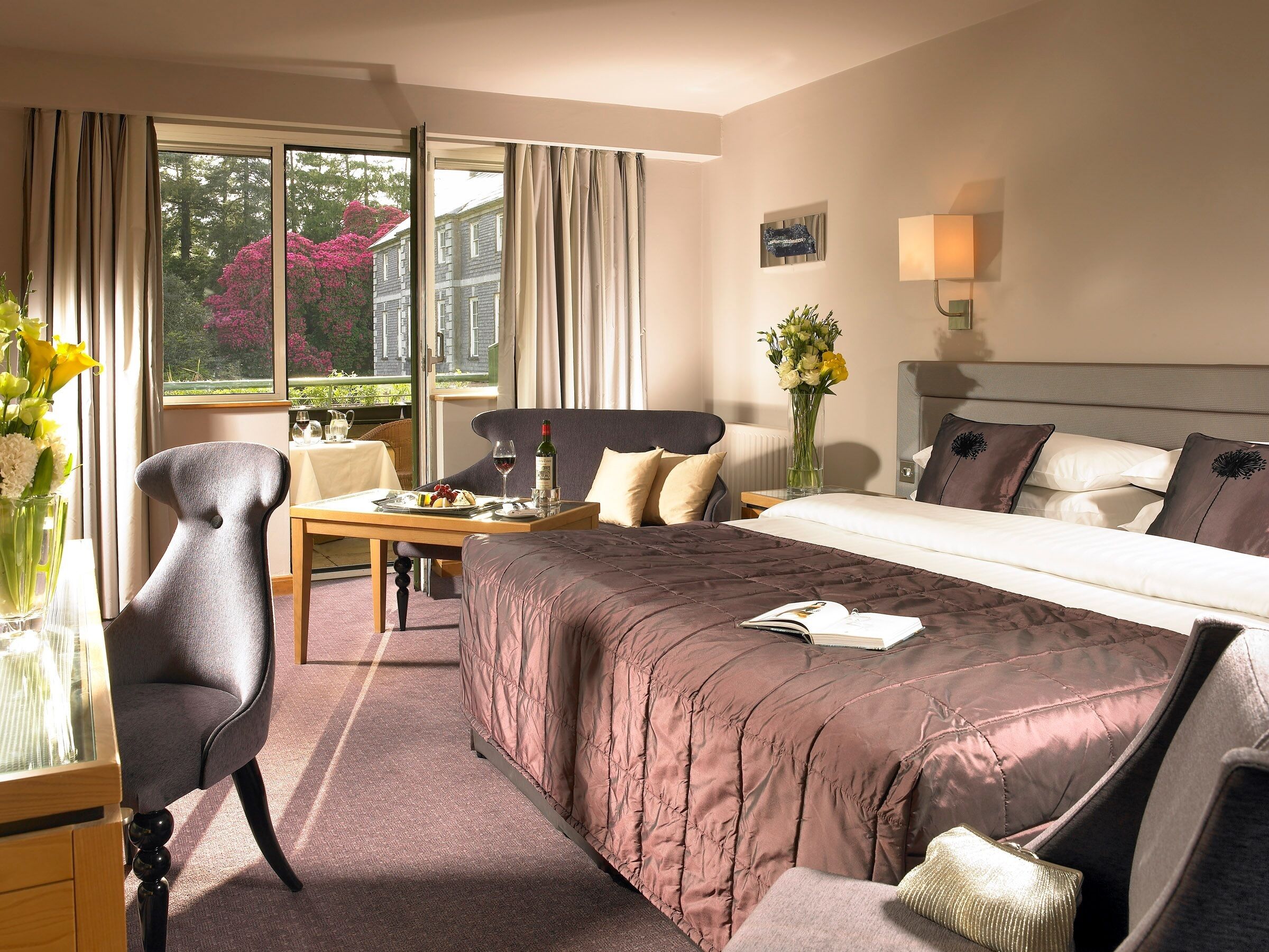 A hotel room with a bed, a desk, a chair, a vase with flowers, and a window with curtains.