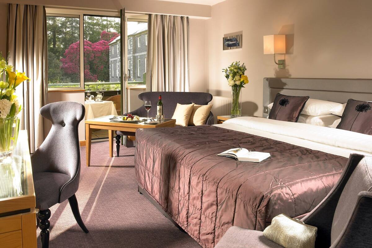 A hotel room with a bed, a desk, a chair, a vase with flowers, and a window with curtains.
