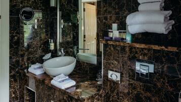 A bathroom with a large white sink, a mirror, and a marble countertop. There are towels stacked on a shelf and a door leading to another room.
