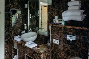 Un baño con un lavamanos grande blanco, un espejo y una encimera de mármol. Hay toallas apiladas en un estante y una puerta que da a otra habitación.