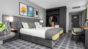 A modern hotel room with a large bed, a desk, and a chair. There are three framed artworks on the wall, a wardrobe, and a mirror. - The Plaza Hotel (Dublin)