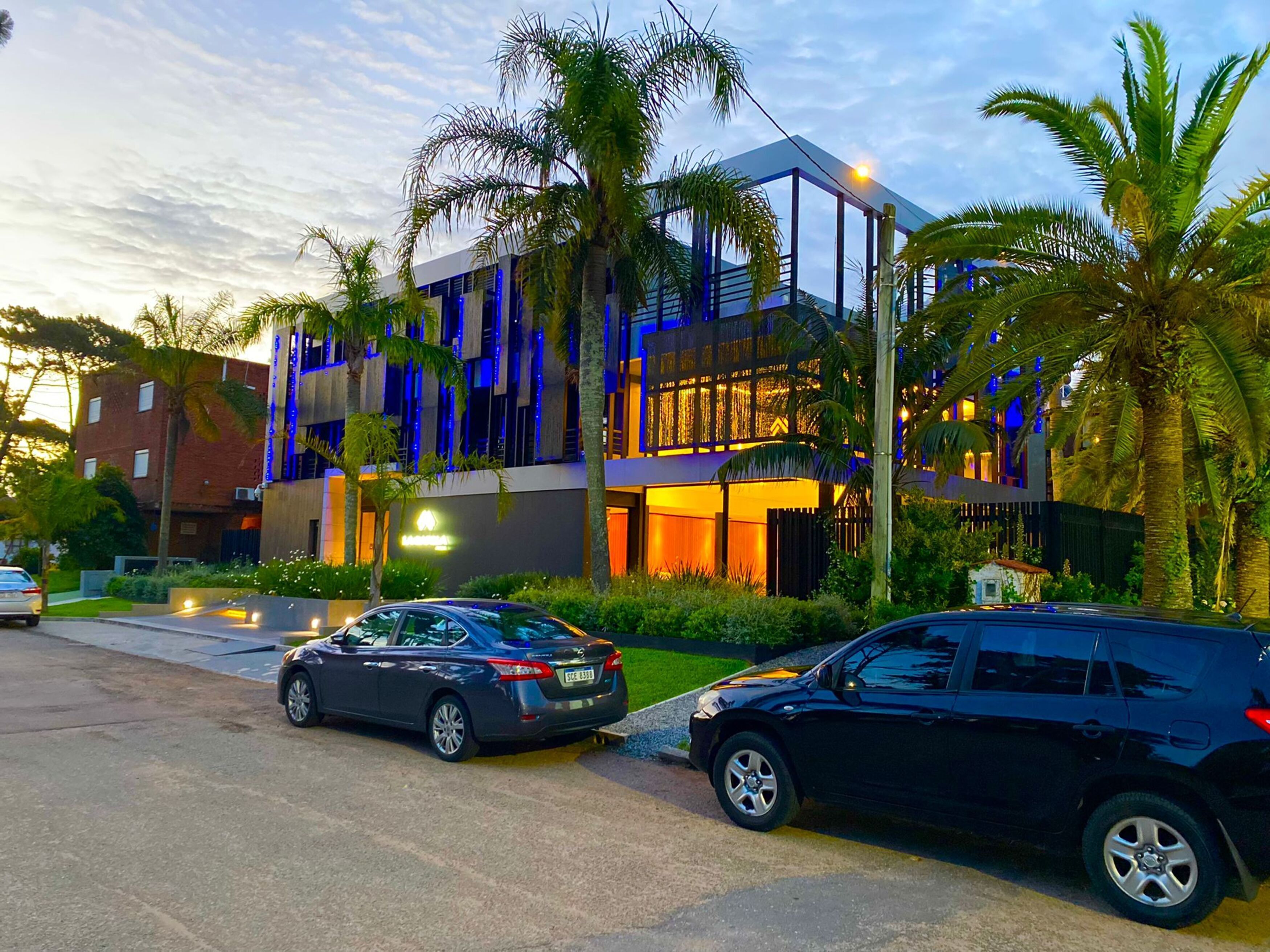 A modern hotel with a glass facade, palm trees, and two cars parked in front.