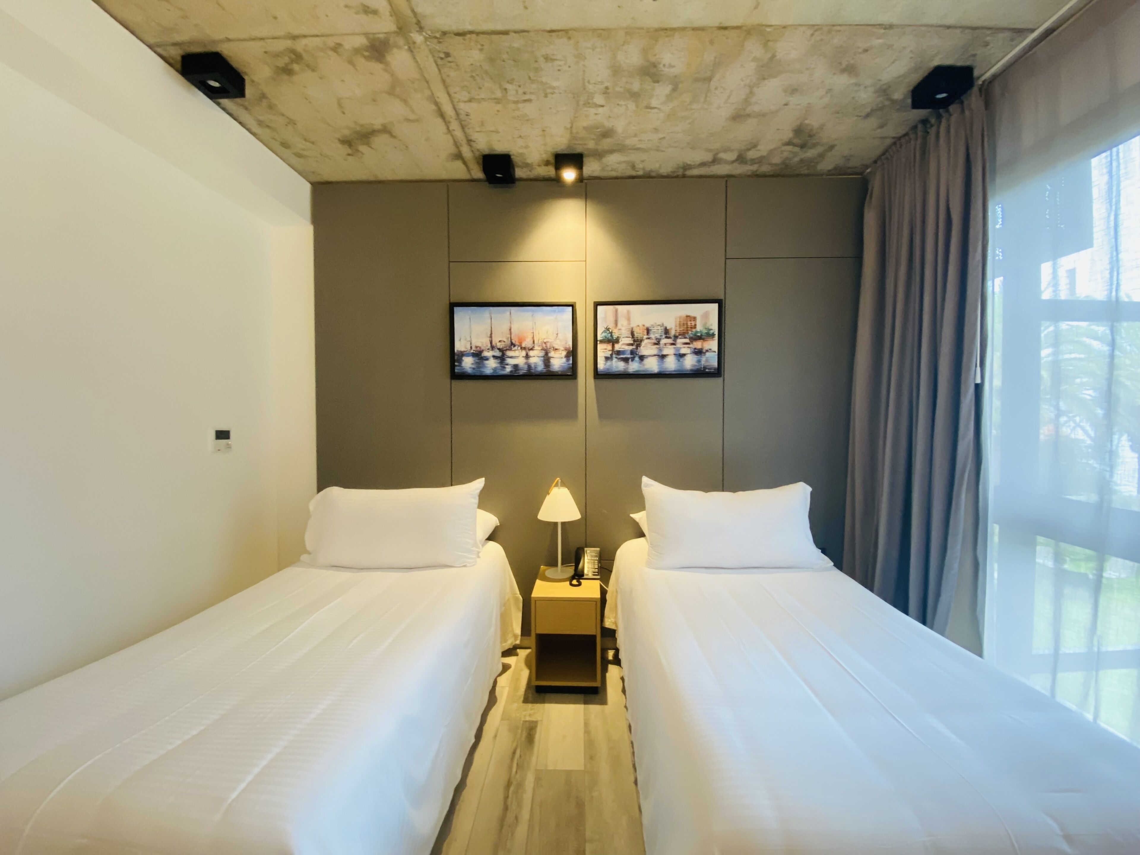 Habitación de hotel con dos camas individuales, una mesita de noche con lámpara y dos cuadros en la pared.