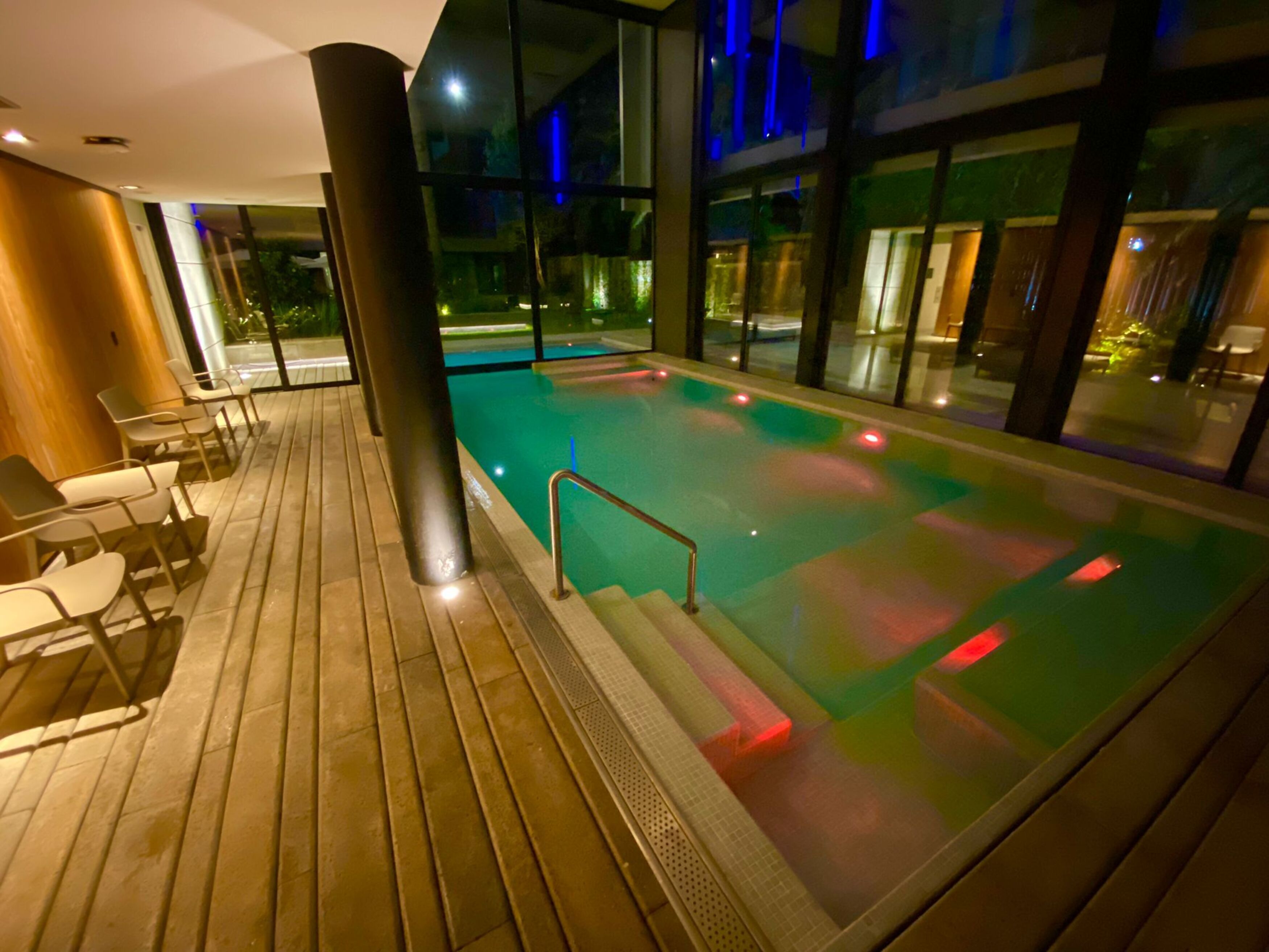 Une piscine intérieure moderne avec une terrasse en bois, un espace salon et de grandes fenêtres.
