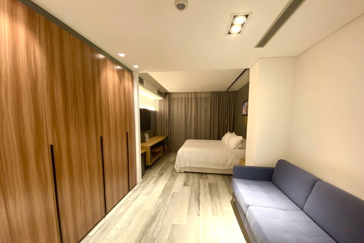 A modern hotel room with a wooden wardrobe, a bed, a blue sofa, and a desk with a chair.