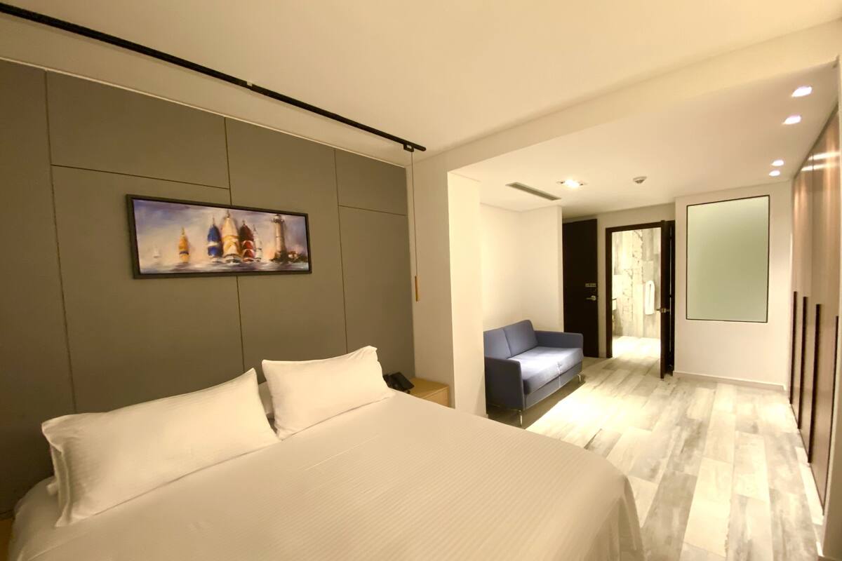 A modern hotel room with a large bed, a blue sofa, a painting of sailboats, and a large mirror.