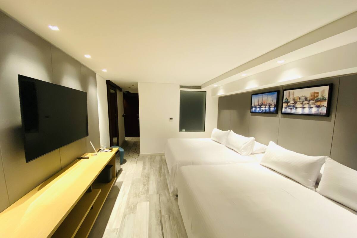 A modern hotel room with a large flat-screen TV, a wooden bench, a bed with white linens, and two framed pictures on the wall.