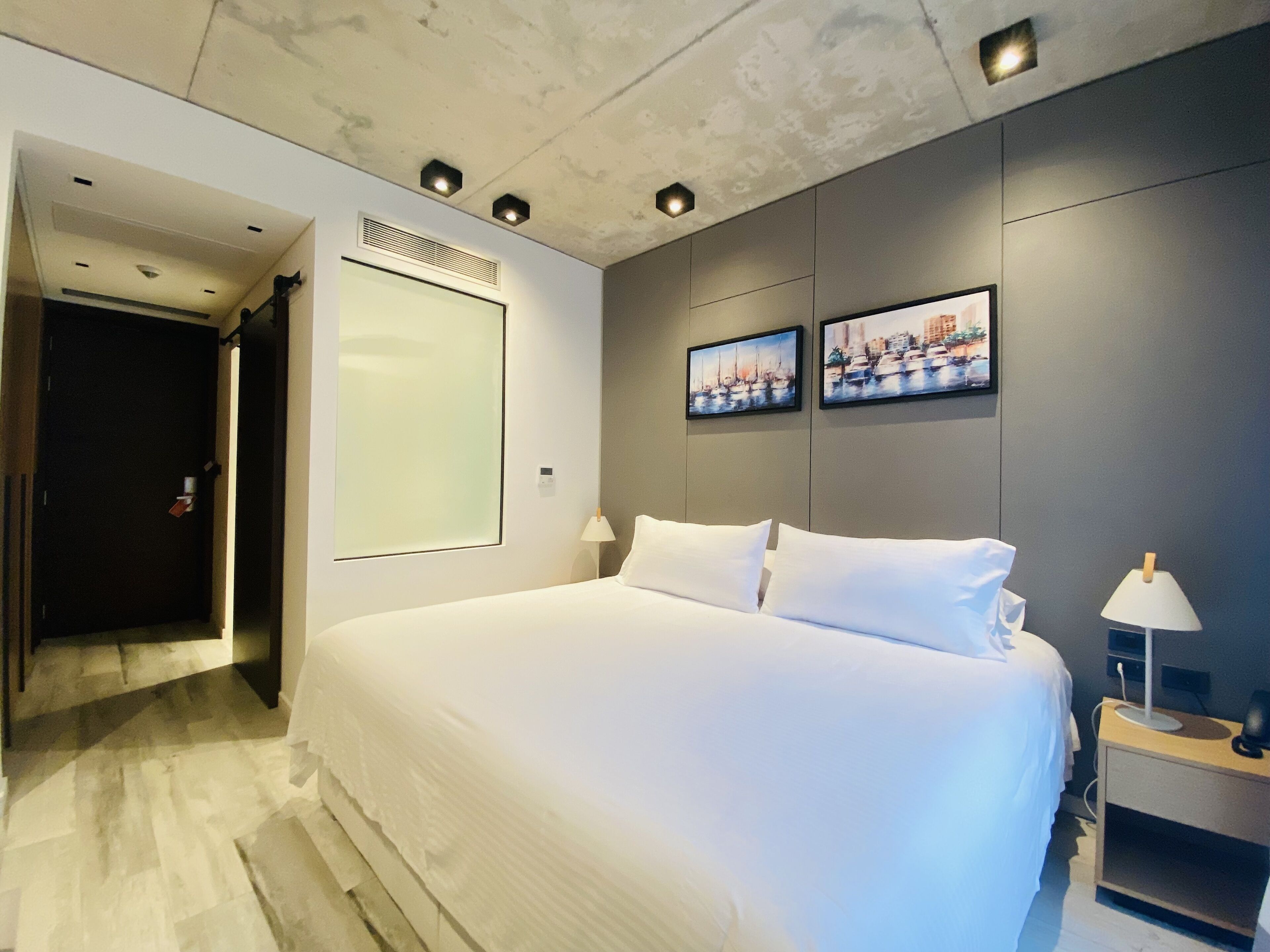 Una habitación de hotel moderna con una cama grande, dos cuadros colgados en la pared, una mesita de noche con una lámpara y una puerta que da a otra habitación.