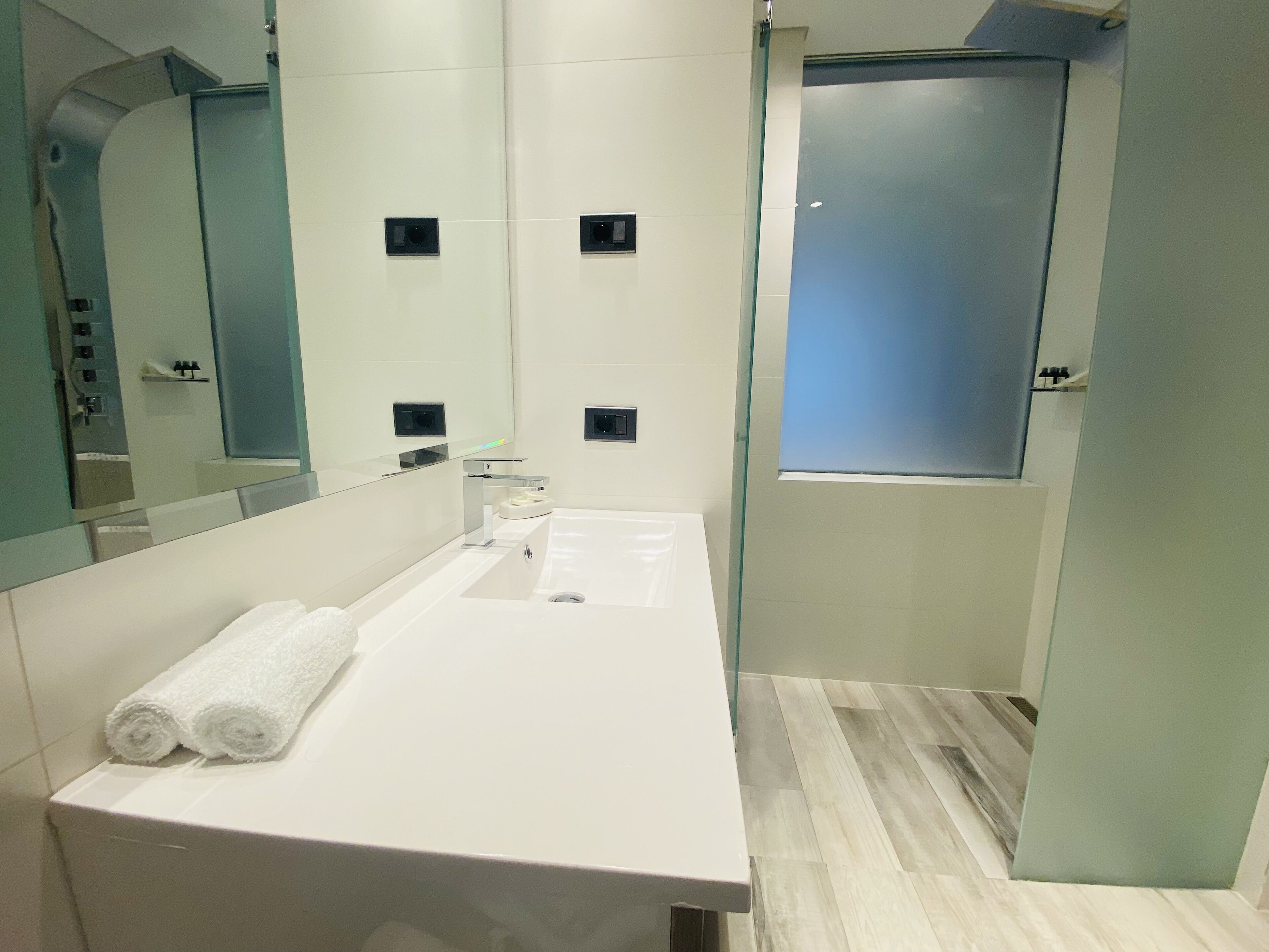 A modern bathroom with a white sink, two towel rolls, a mirror, and a glass partition.