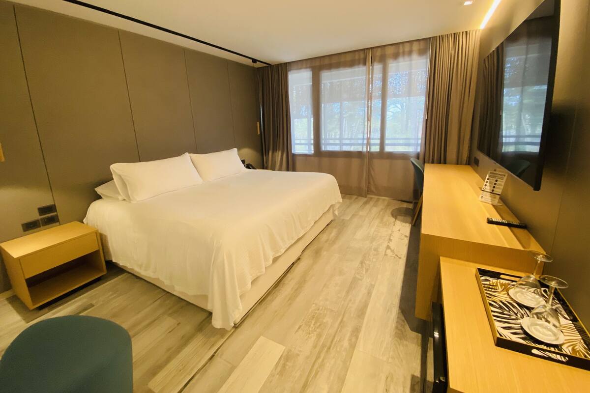 A hotel room with a large bed, a desk with a flat-screen TV, a wooden nightstand, and a seating area.