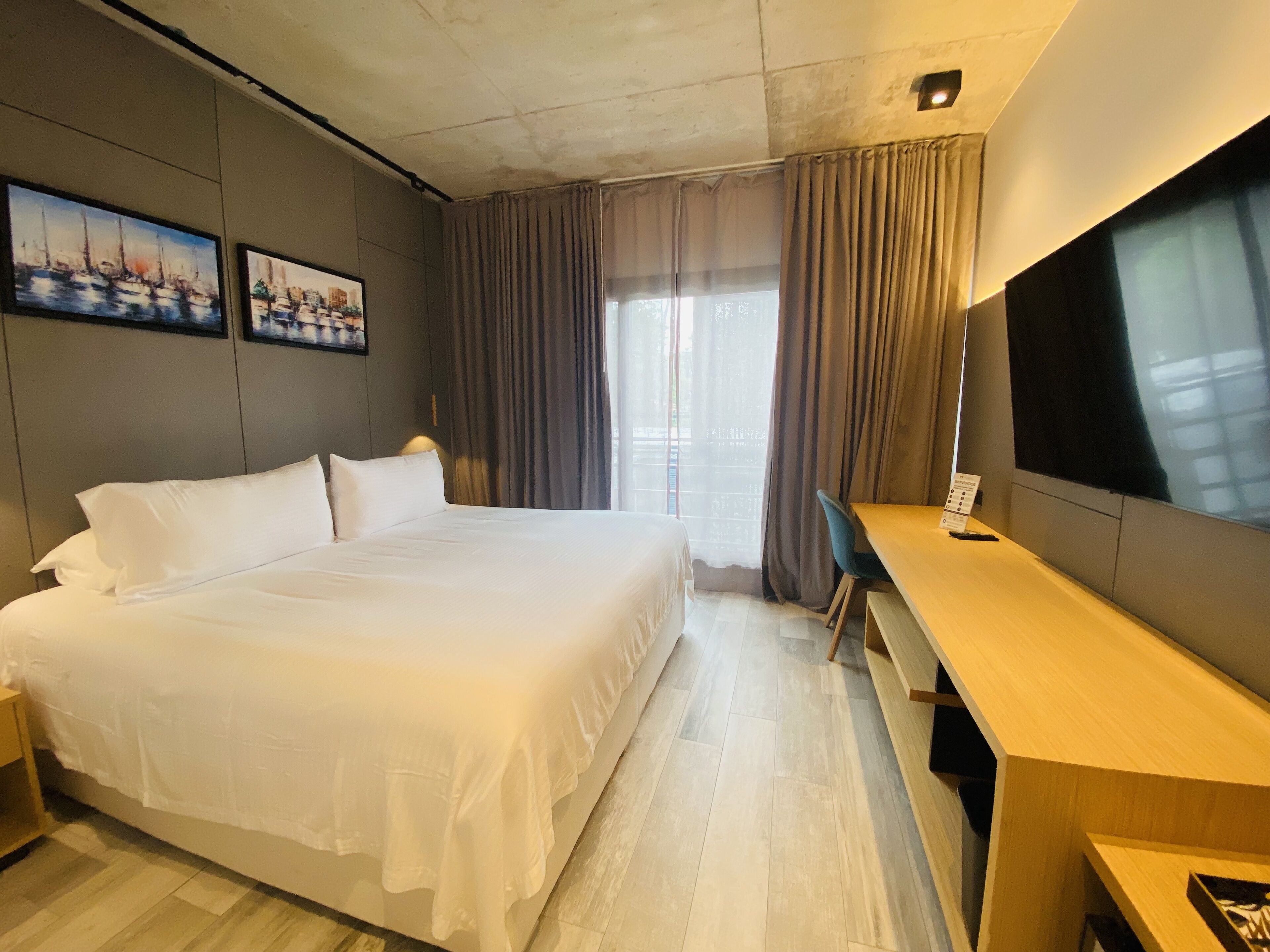 A hotel room with a large bed, a desk, a TV, and two paintings on the wall.