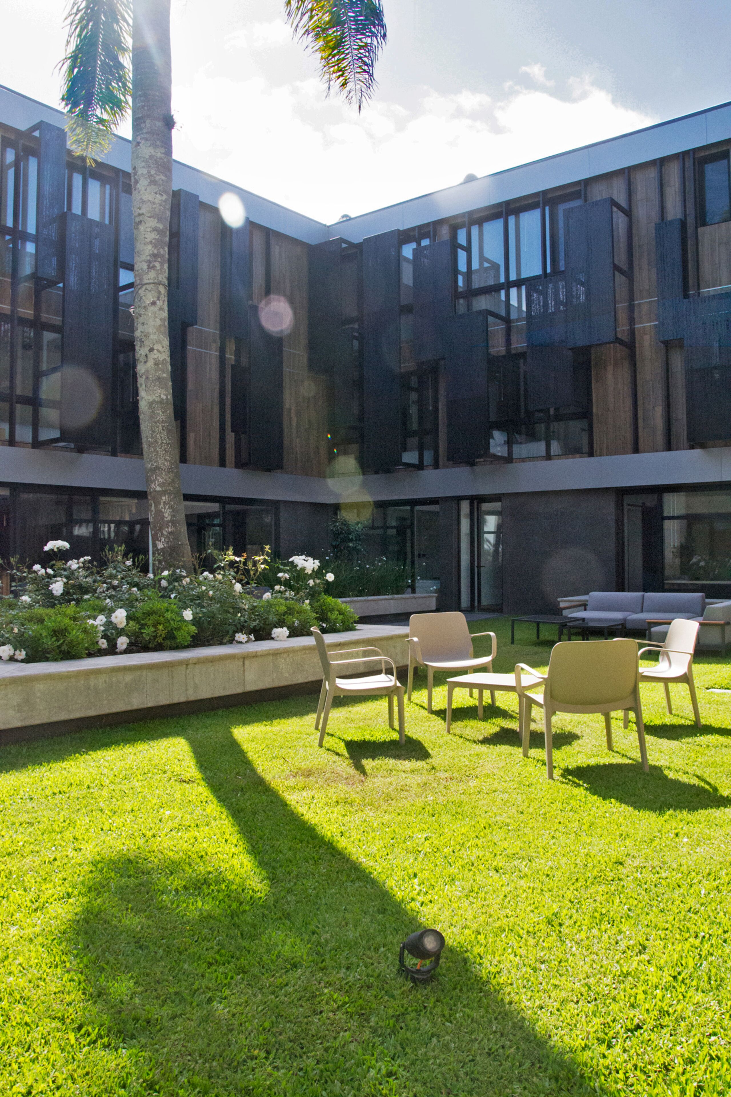 A modern building with a courtyard featuring a lawn, seating area, and a palm tree.