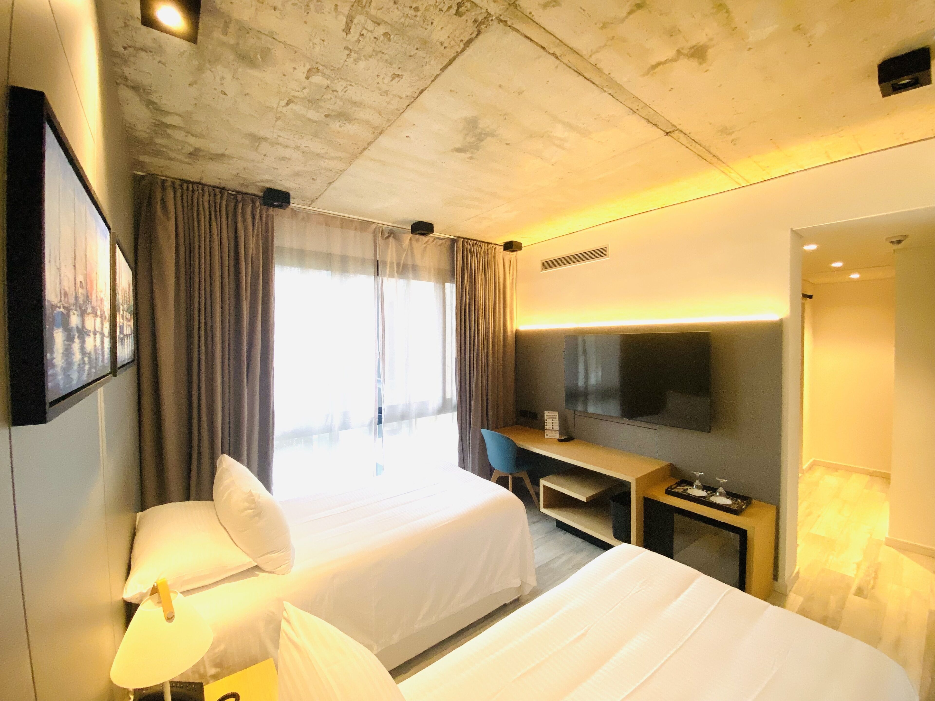 Quarto de hotel com duas camas, uma mesa, uma televisão e uma janela grande com cortinas.
