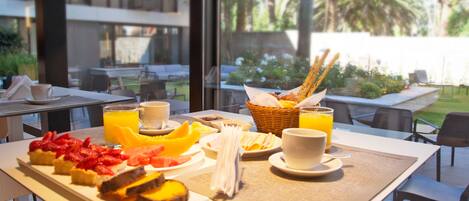 Mesa de desayuno con fruta, pasteles y jugo, con vista a un jardín con palmeras.