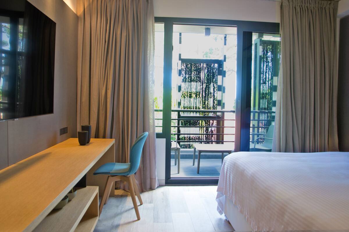 A hotel room with a bed, a desk, a chair, and a balcony with a view of greenery.