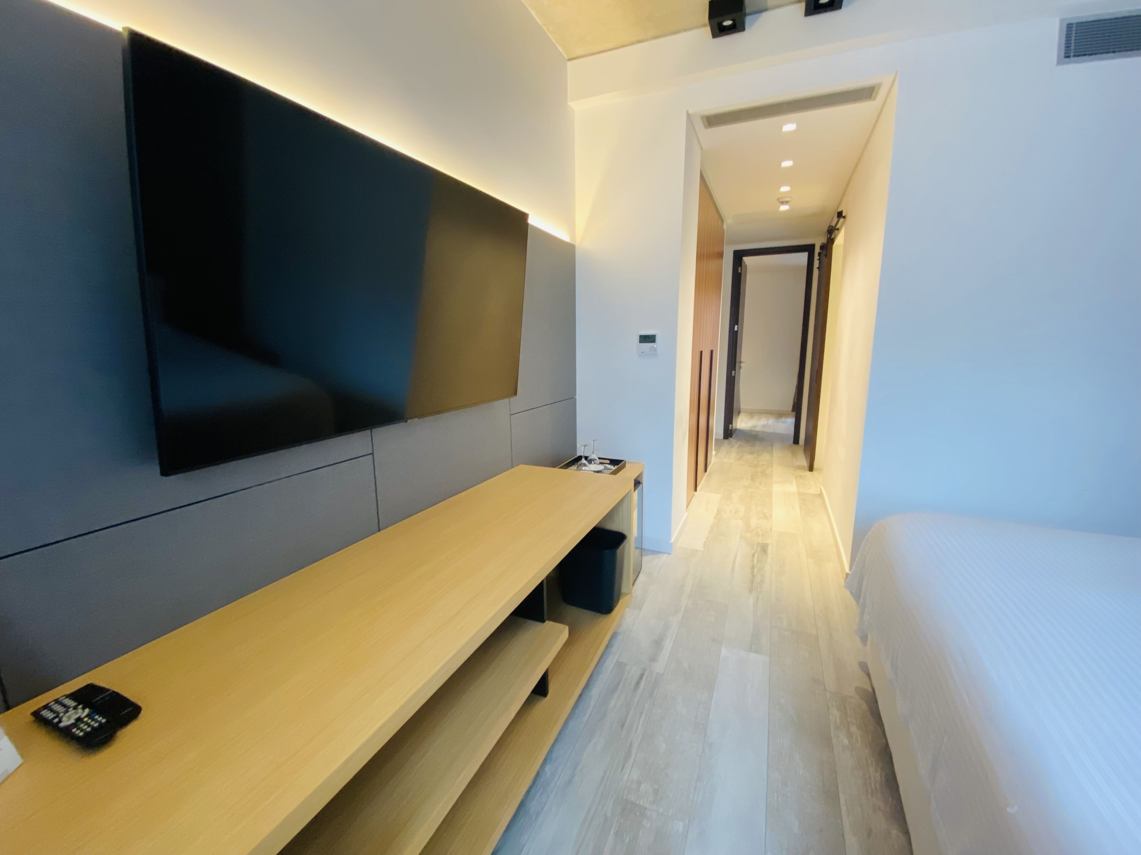 Quarto de hotel moderno com televisão de tela plana na parede, um banco de madeira com controle remoto, uma cama e um corredor que dá para outro quarto.