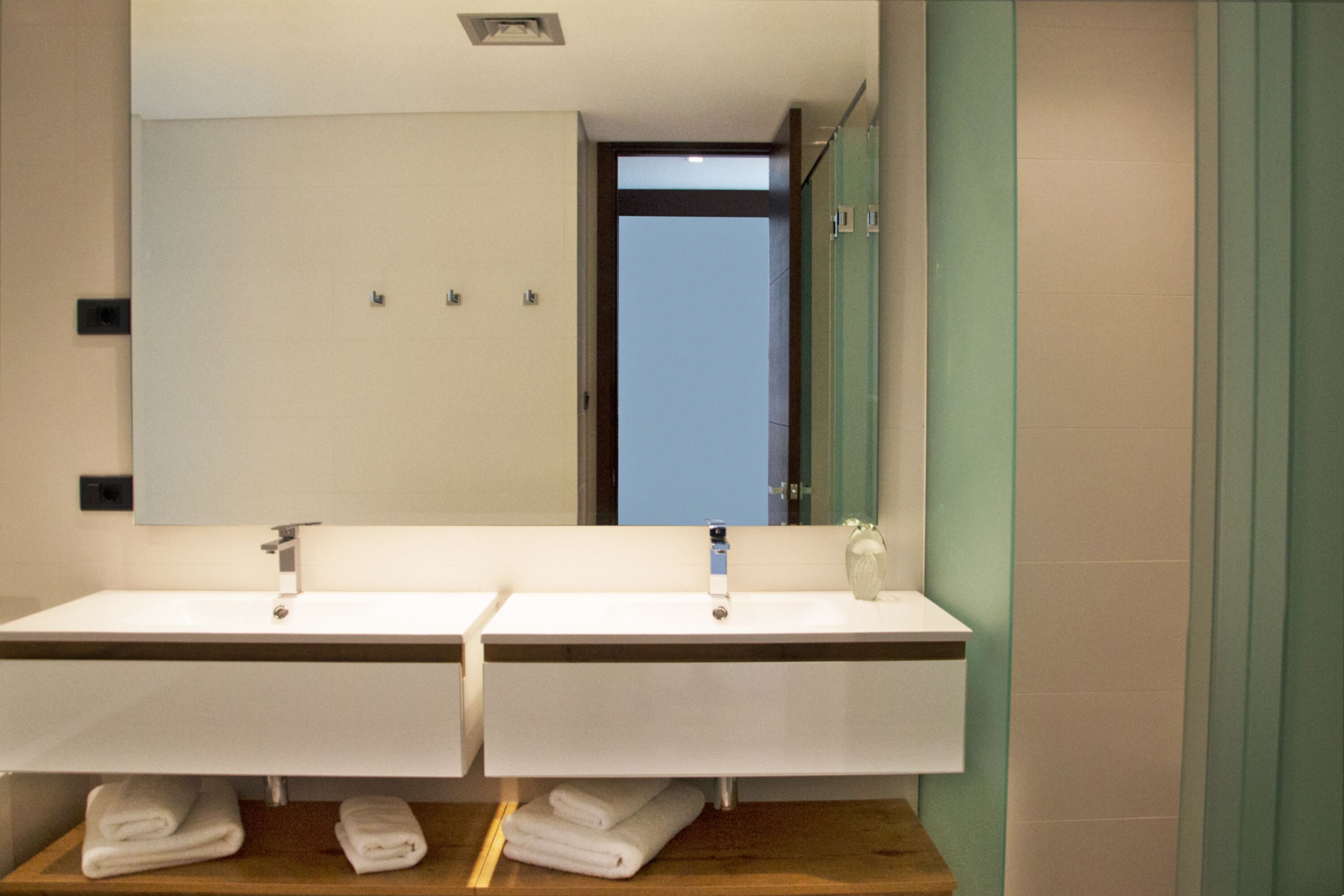 Un baño moderno con dos lavamanos, un espejo grande y vistas al cielo a través de una puerta abierta.