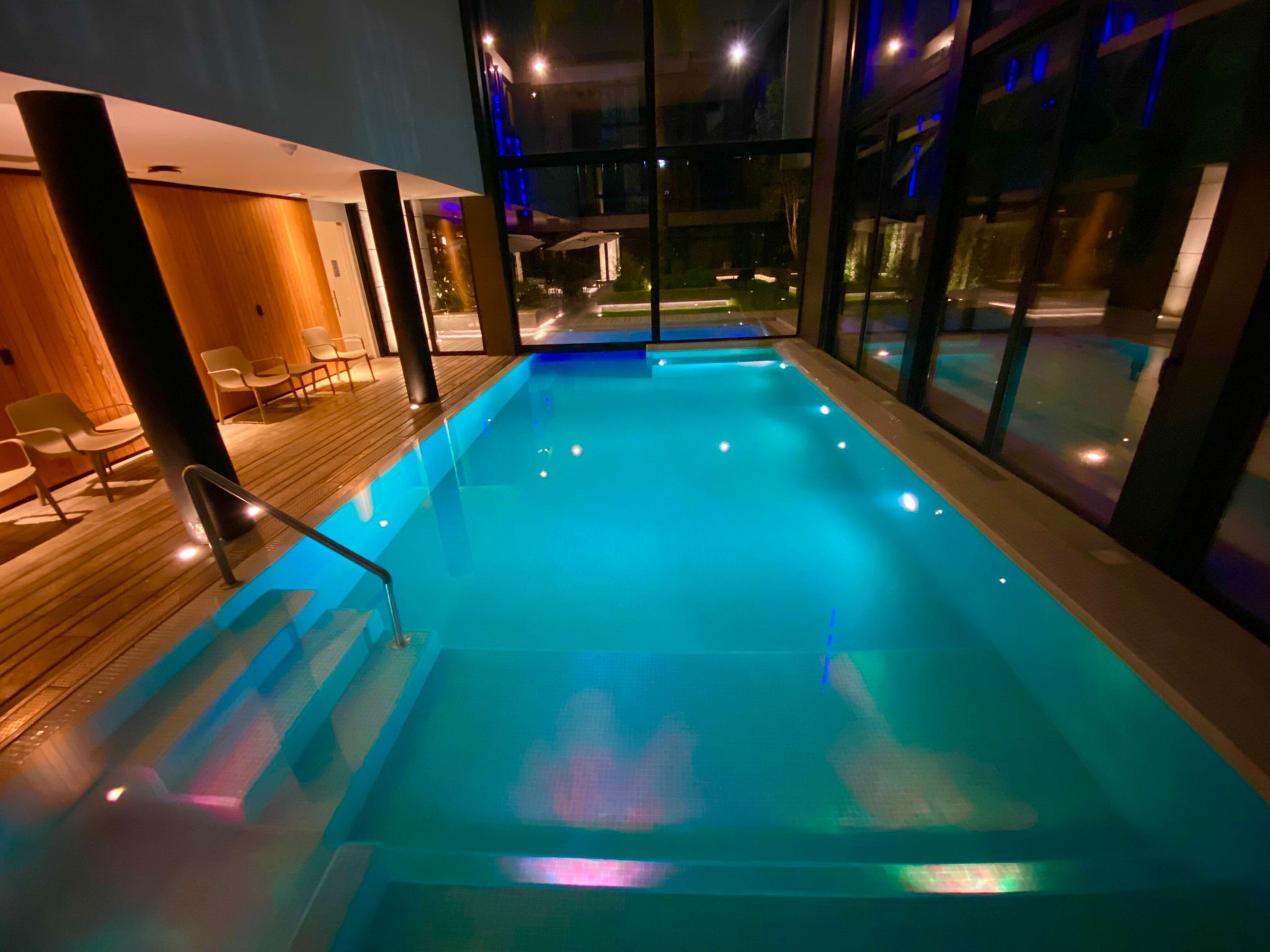 An indoor swimming pool with clear blue water, wooden deck, and glass walls offering views of the outside.