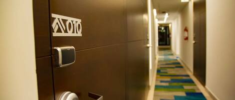 Un couloir d’hôtel moderne avec une porte métallique, un logo bien visible et un sol recouvert d’une moquette colorée.
