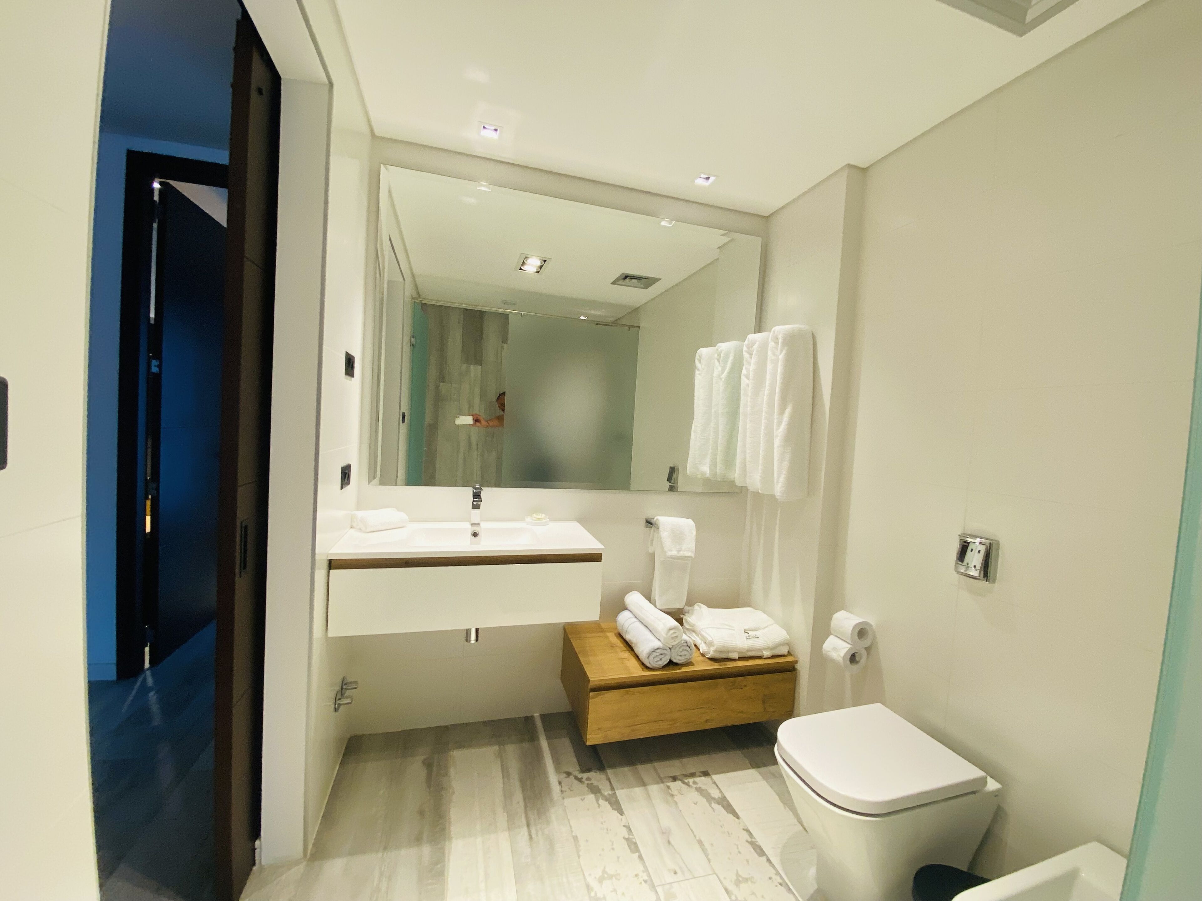 Ein modernes Badezimmer mit einem weißen Waschbecken, einem Spiegel und einer Toilette. Eine Holzbank mit Handtüchern und einem Handtuchhalter an der Wand.