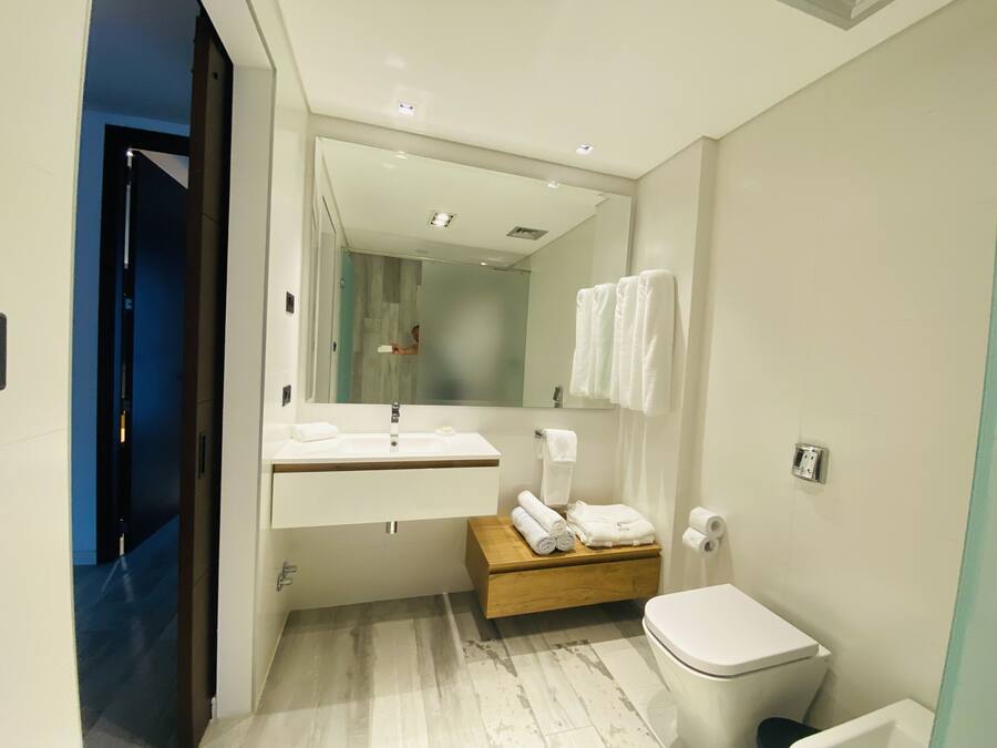 Un baño moderno con lavamanos, espejo y retrete blancos. Hay un banco de madera con toallas y un perchero en la pared.