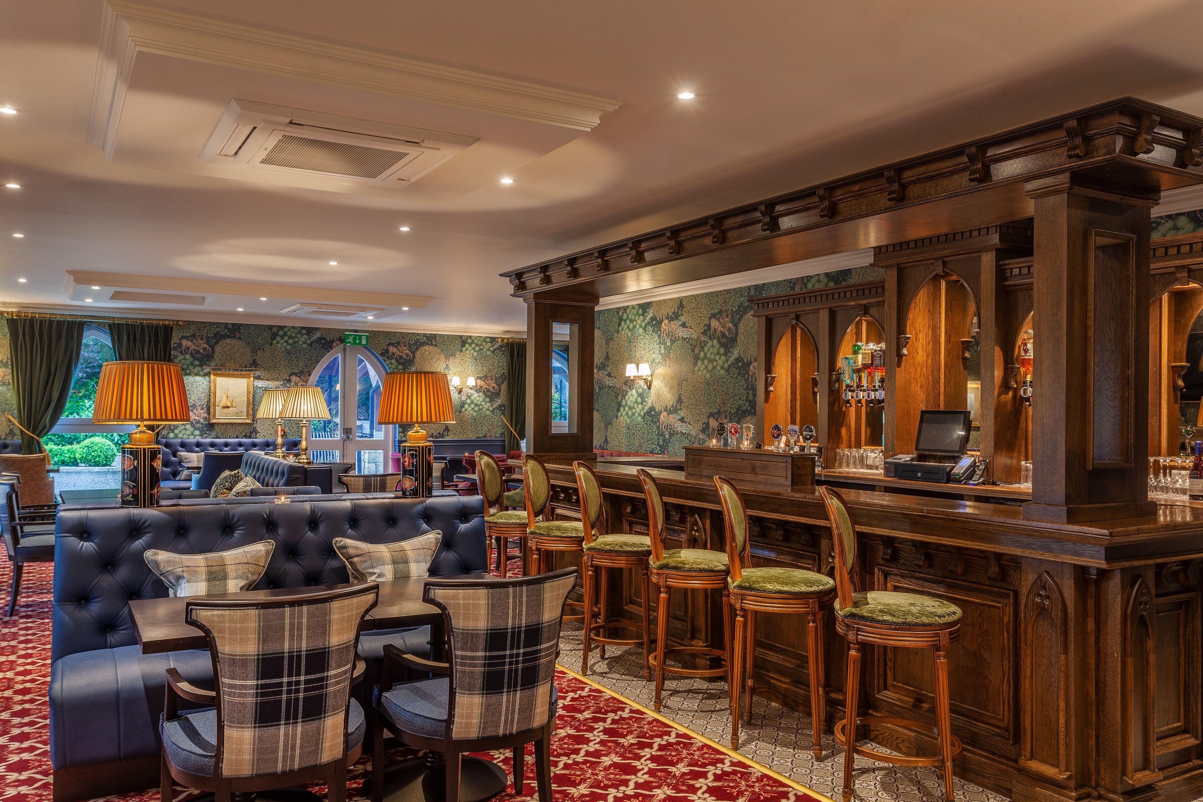 Zona de bar con mostrador de madera oscura, varios taburetes y un área de estar confortable con sofá capitonado y sillas a cuadros.