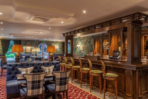 Zona de bar con mostrador de madera oscura, varios taburetes y un área de estar confortable con sofá capitonado y sillas a cuadros.
