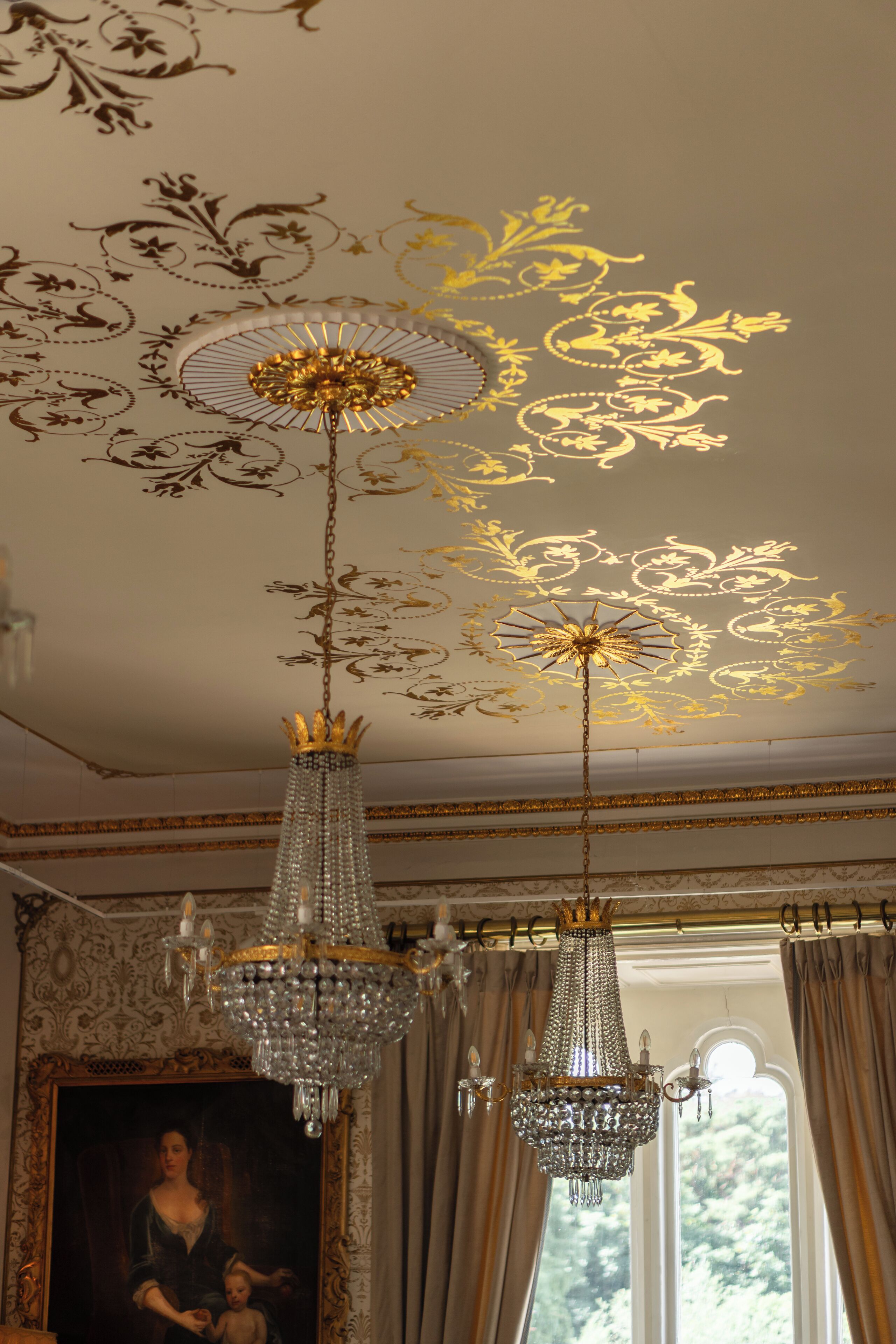 A chandelier with a gold-colored center and crystal elements, hanging in a room with ornate ceiling designs and a large window with curtains.