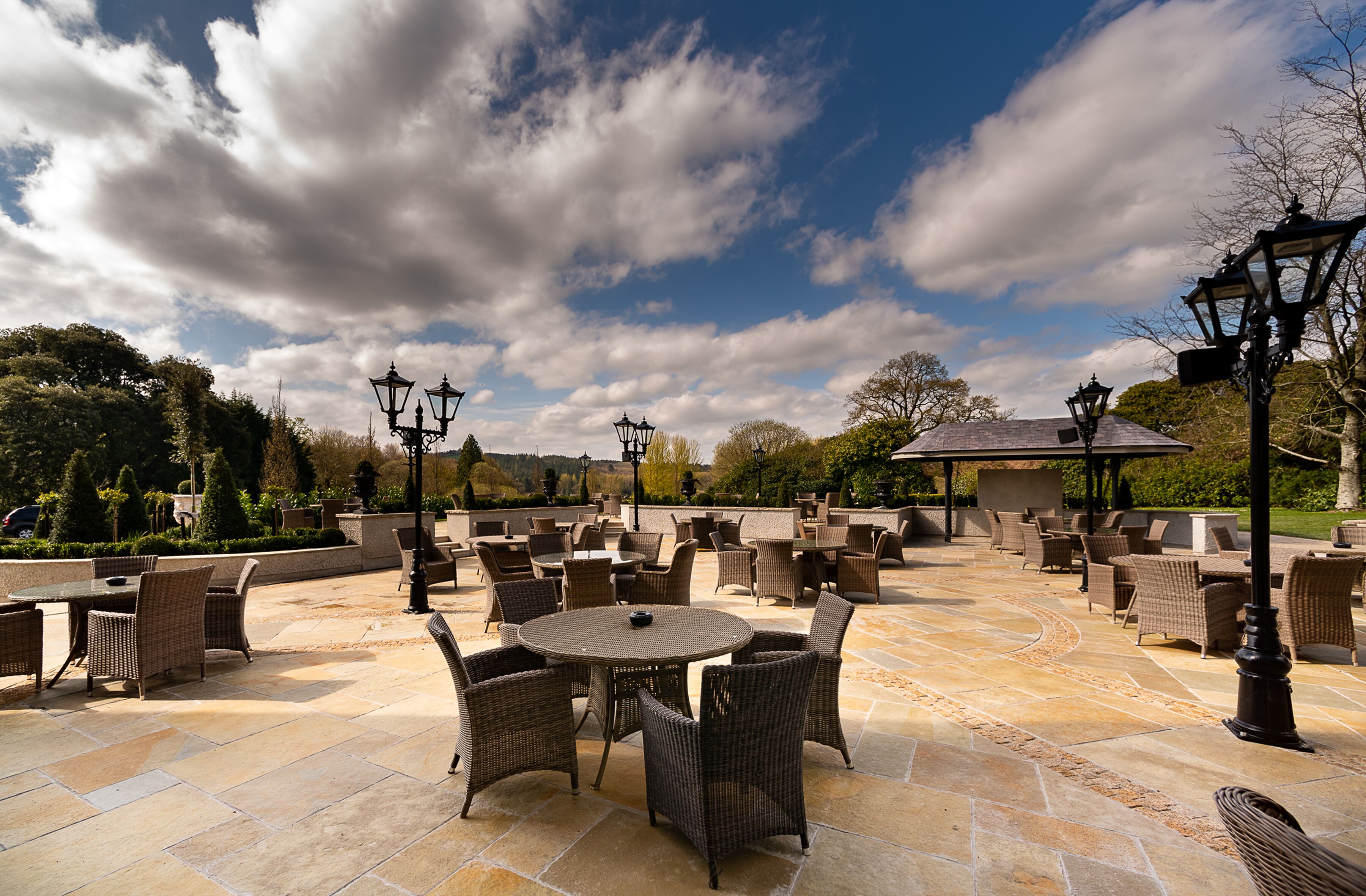 An outdoor patio with wicker furniture and stone flooring, surrounded by trees and a clear sky.