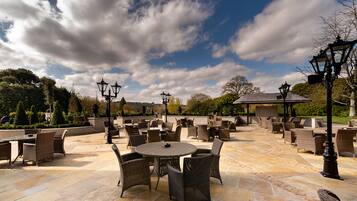 An outdoor patio with wicker furniture and stone flooring, surrounded by trees and a clear sky.
