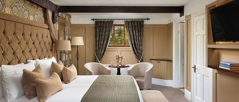 A hotel room with a large bed, two armchairs, a small table with a lamp, and a TV mounted on the wall.
