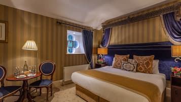 Una habitación de hotel con una cama grande, una mesa redonda con sillas, una ventana con cortinas y una mesita de noche con lámpara.